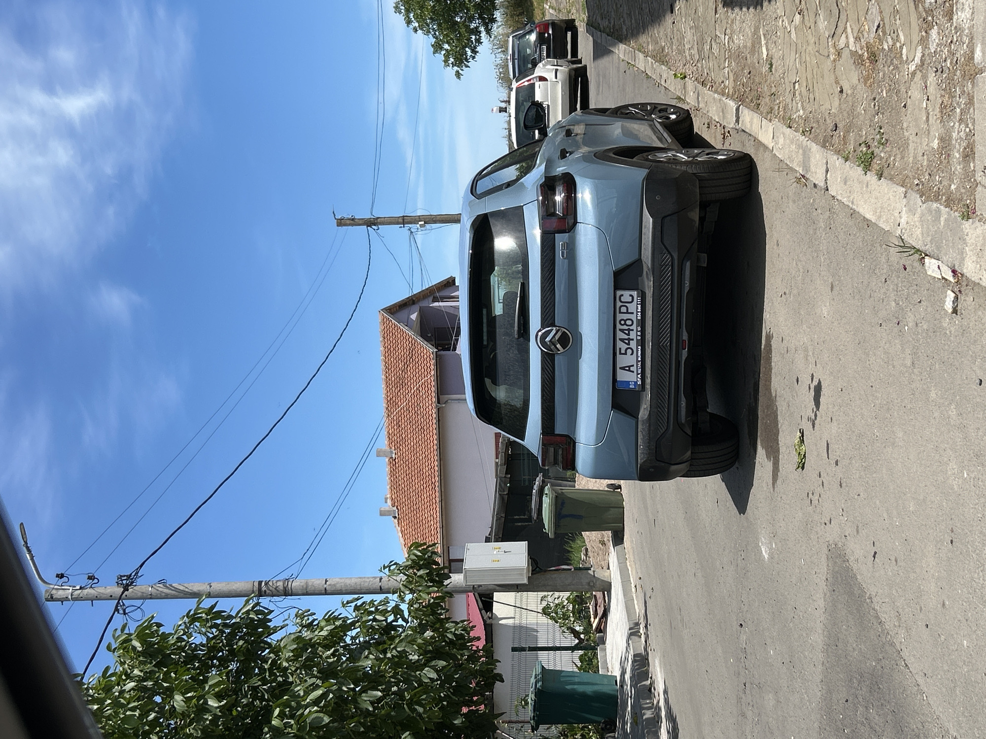 In Aheloy, Bulgaria, on a sunny day with a clear blue sky and scattered clouds, a light blue Citroen C4 is parked on an asphalt street next to a stone curb. The car, facing away from the camera, displays a license plate reading "BG A 5448 PC". Behind the Citroen, a white Volvo station wagon and a dark-colored sedan are also parked on the street. On the left side of the street, a green recycling bin stands near a fence and a building with a red tiled roof. Further down the street, another green bin is visible. A concrete utility pole with electrical wires runs along the side of the road. The environment appears to be a residential area with houses and vegetation.