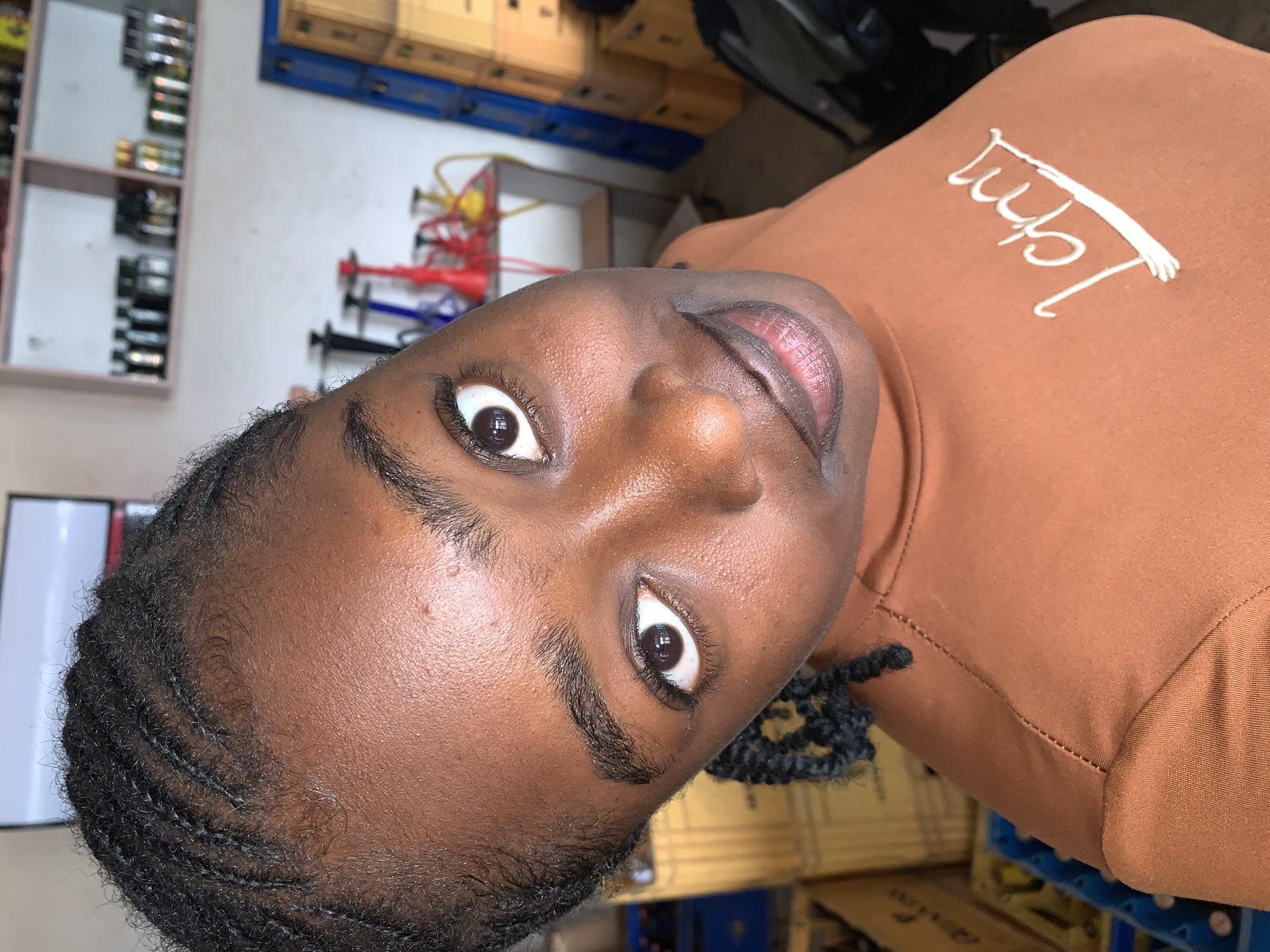 A young woman with a dark complexion, featuring small, tight black cornrows and visible baby hairs at the hairline, is depicted in a close-up, upward-angled shot. Her wide, dark brown eyes look directly towards the camera, and her lips are closed in a subtle, engaging smile. Her skin shows a natural sheen on the forehead and cheeks. She is dressed in a close-fitting, rust-brown colored top with a high neck, which has a white, stylized script, possibly reading "I am," embroidered or printed on the left chest.
The background indicates an indoor commercial or storage environment within New Stadium, Nigeria. In the upper left, a white rectangular screen or television is partially visible. The upper right segment displays two light-colored wall-mounted shelves holding multiple small bottles and containers. Below the shelves, an arrangement of various colored hookahs, including black, blue, red, and yellow, is visible on a surface. The lower portions of the background are occupied by stacks of brown cardboard boxes and blue plastic crates, consistent with inventory or merchandise storage. The walls appear to be plain and light-colored.
