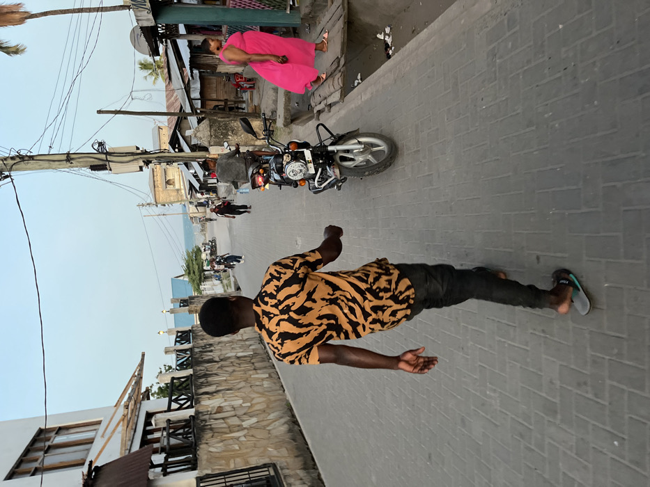 The photograph is rotated 90 degrees clockwise, depicting a lively street scene in what appears to be a coastal town, likely Bagamoyo, Tanzania.
In the foreground, centered, a man with dark skin walks away from the viewer, looking towards the right side of the frame. He wears a short-sleeved shirt with a distinctive black and orange tiger-stripe pattern and dark trousers. His left foot is bare, while his right foot is in a blue and white flip-flop with the word "AFRICA" visibly printed on its strap.
Behind him, on the same paved street made of grey interlocking bricks, a black motorcycle is parked. Further down the street on the left, a woman in a vibrant fuchsia-pink sleeveless dress stands near a building, facing left. Several other figures are visible in the distance, suggesting street activity.
The street is lined with buildings of varying construction. On the left, a low stone wall with wooden railings fronts a building with a white facade and dark wooden elements. On the right, older structures with corrugated metal roofs and stone or concrete walls are present. Tall utility poles with numerous tangled wires run along the right side of the street. Palm trees are scattered throughout the scene.
In the far background, under a clear, pale blue sky, the calm waters of the ocean are visible, suggesting the town is situated directly on the coast. The lighting indicates it is daytime, possibly late afternoon, with soft, even illumination. The scene conveys the atmosphere of a typical, bustling local street in a warm, coastal environment.