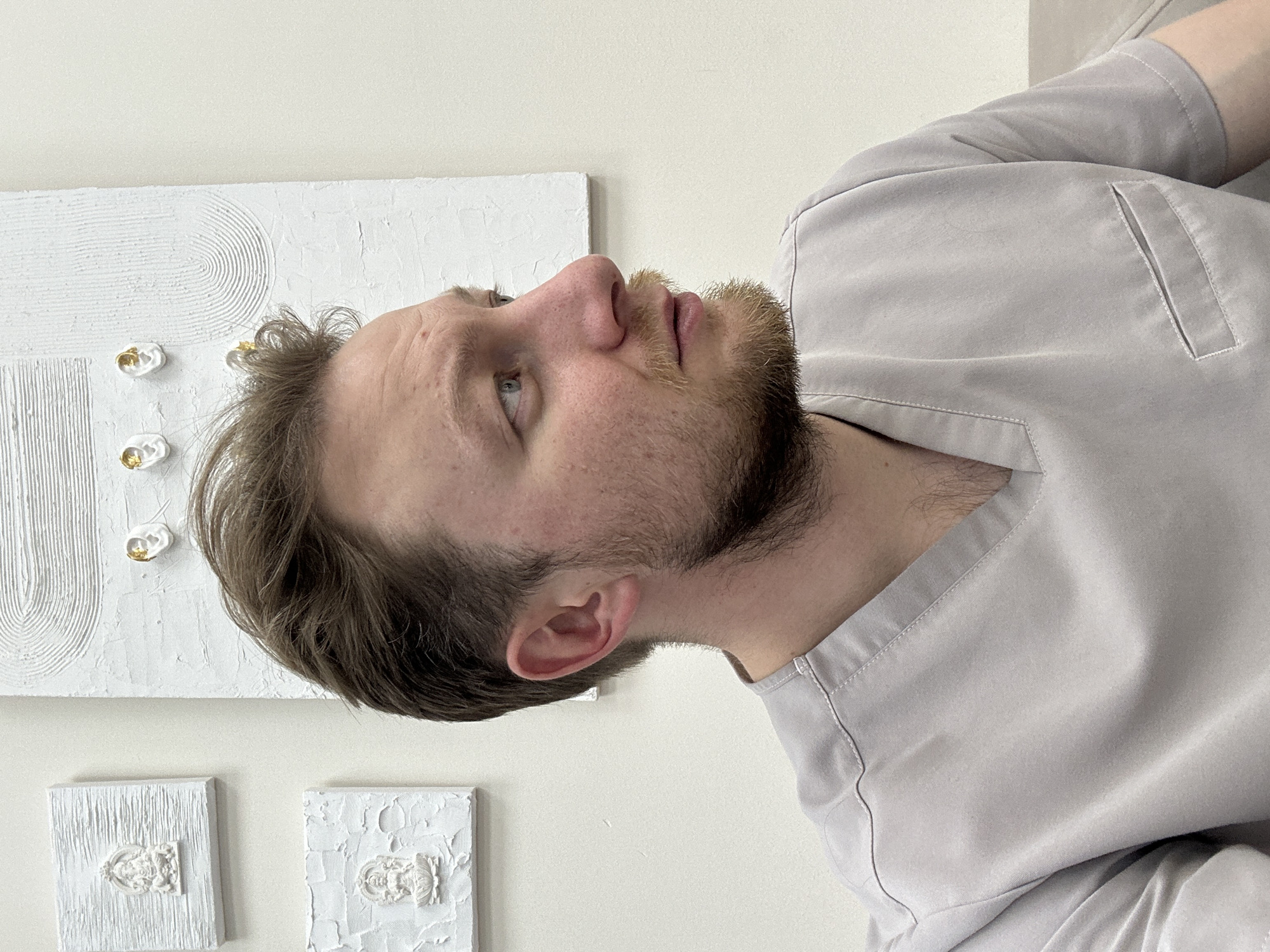 A fair-skinned man with short brown hair and a beard is viewed from the side and slightly below, his gaze directed upwards. He wears a light gray or lavender collared shirt, possibly scrubs. Behind him is a white wall adorned with three pieces of textured white artwork. The city of Sochi, Russia, cannot be determined from the image.