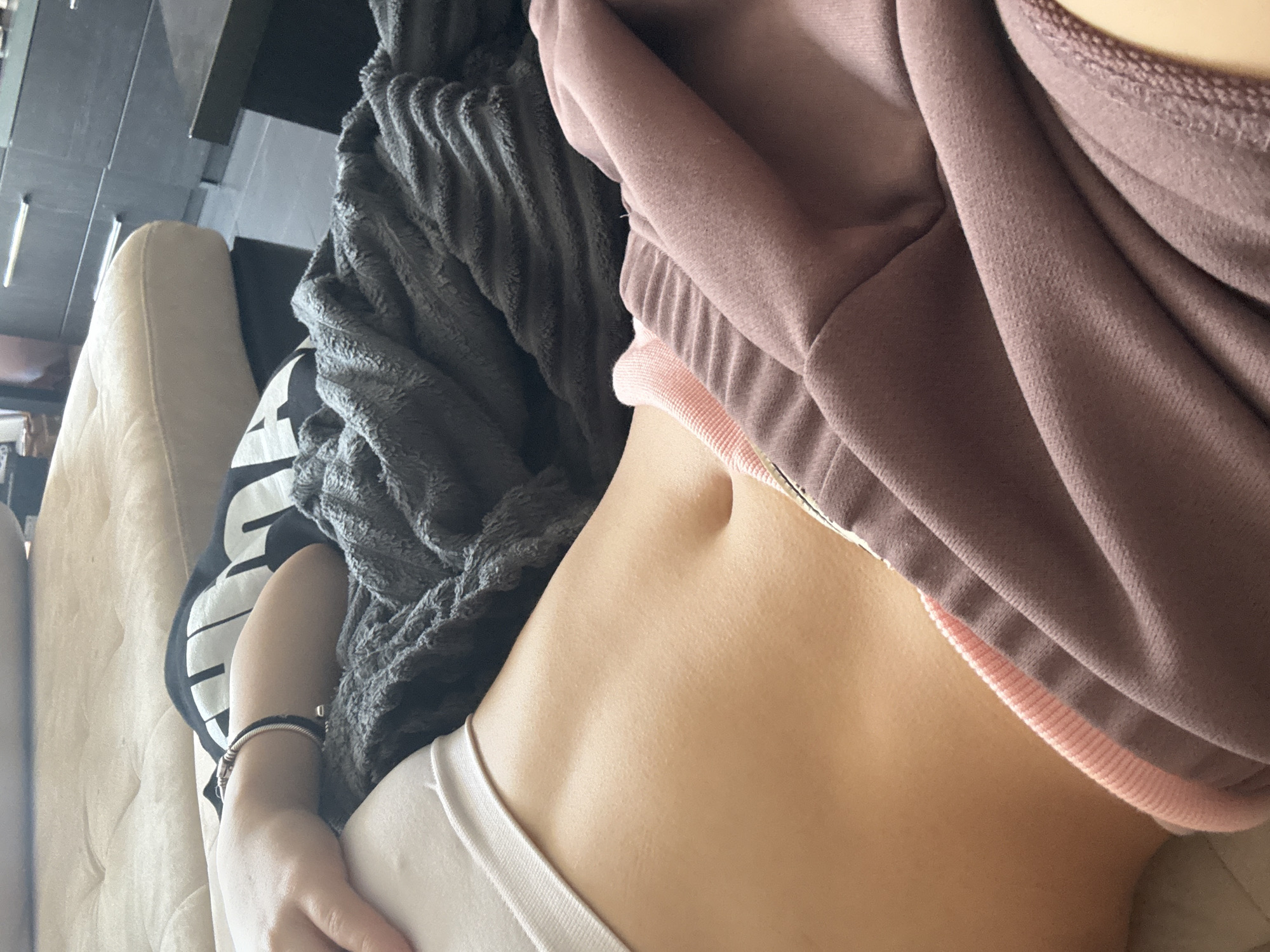 A close-up, overhead shot focuses on a person's torso, specifically their toned abdomen and midriff. They are lying on a light-colored couch. A dark grey blanket is draped over the lower part of their torso, partially obscuring some of their clothing. Their right arm, adorned with a silver bracelet, rests across the blanket. A hint of a black t-shirt with white lettering is visible beneath the blanket. The person is wearing a white bra and pink-trimmed sweatpants. The background shows a dark, polished surface, likely a table, and a dark dresser with silver handles, suggesting an indoor setting, possibly a living room. There is no information in the image to indicate the location is in the city of Sofia, Bulgaria.