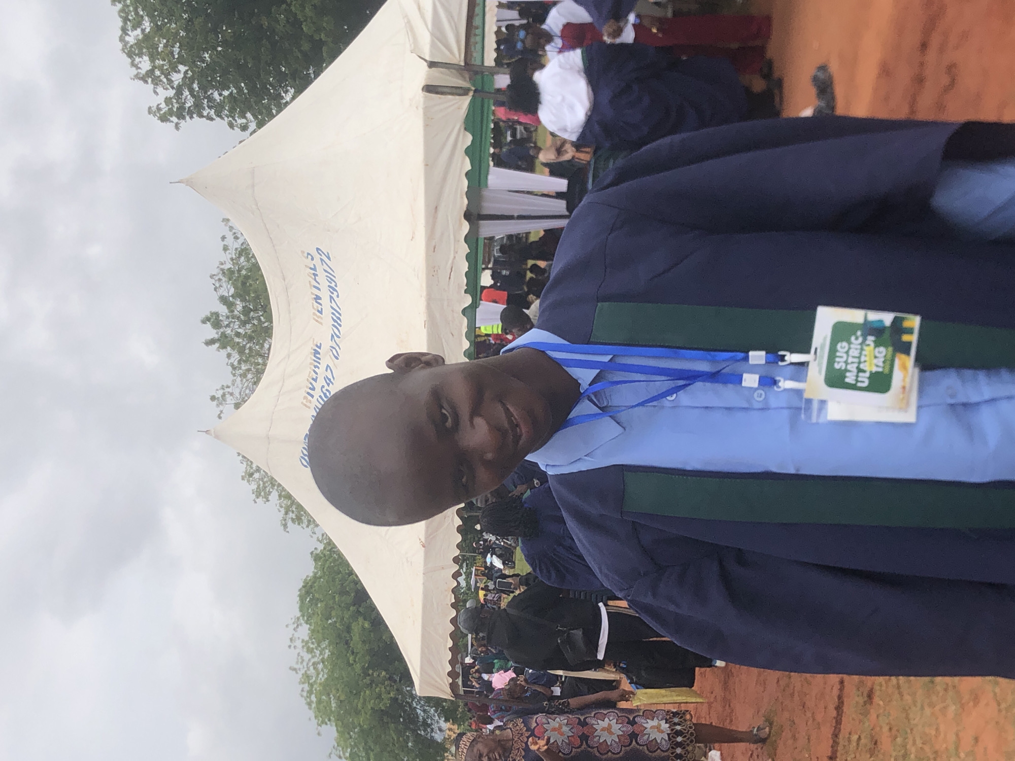 A medium shot depicts a person in what appears to be a graduation gown, wearing a tag labeled "SUG MATRICULATION TAG," positioned in front of a large tent. The tent has the words "RIVERLINE RENTALS 07081799172, 07047110647" printed on it. The setting seems to be outdoors, with a crowd of people and trees visible in the background. The location is likely Nsugbe, Nigeria, based on the information present on the tag.