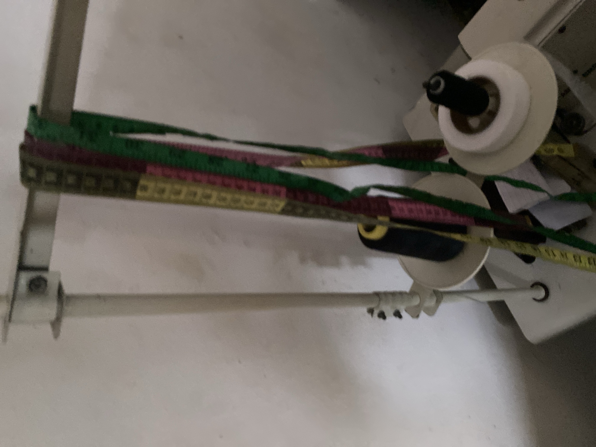The image shows a close-up of a sewing machine with spools of thread and measuring tapes. The machine appears to be part of an industrial sewing setup, with multiple spools arranged on a spindle. The measuring tapes, in various colors like green, pink, and yellow, are draped across the spindle and appear to be ready for use in a sewing project. In the background, a white wall serves as the backdrop, and a hint of the machine's metallic structure is visible. The lighting is somewhat dim, suggesting an indoor setting. There are no people visible in the frame, and the focus is entirely on the sewing equipment. The image is taken from a slightly elevated angle, looking down at the spindle and tapes. The location context of Jalingo, Nigeria, is provided but not visually evident within the image itself.