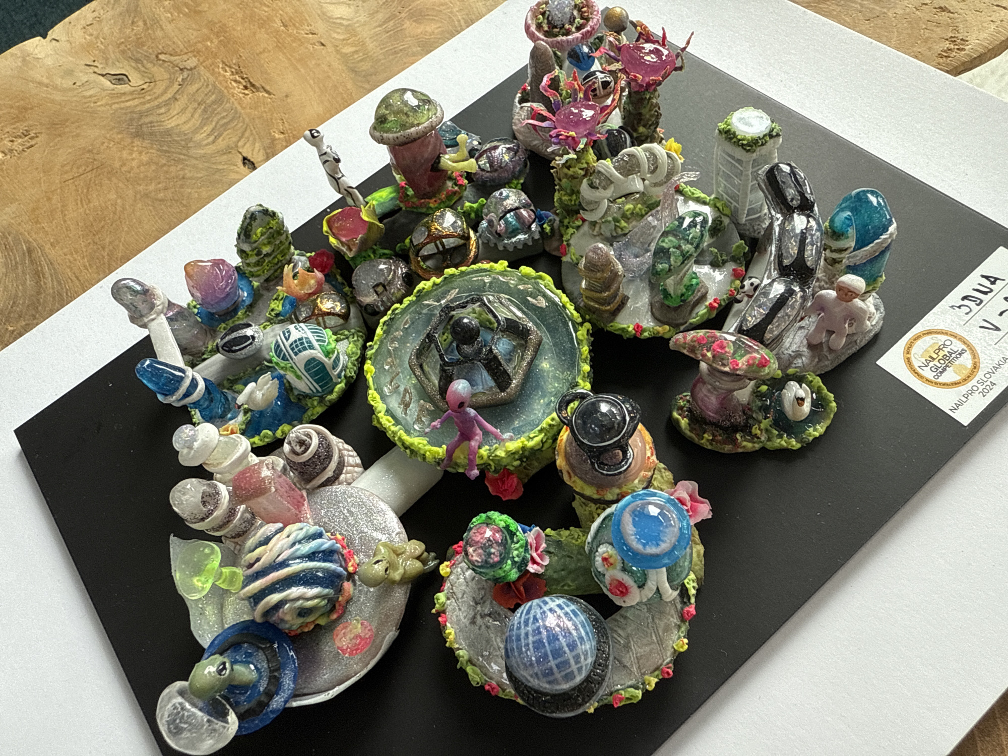 A collection of miniature, fantasy-themed objects sits on a black and white display platform. The objects are colorful and intricately designed, resembling a mix of otherworldly landscapes and structures. The platform is labeled "NAILPRO GLOBAL COMPETITIONS SLOVAKIA 2024". There is no verifiable information in the image about Burgas, Bulgaria.