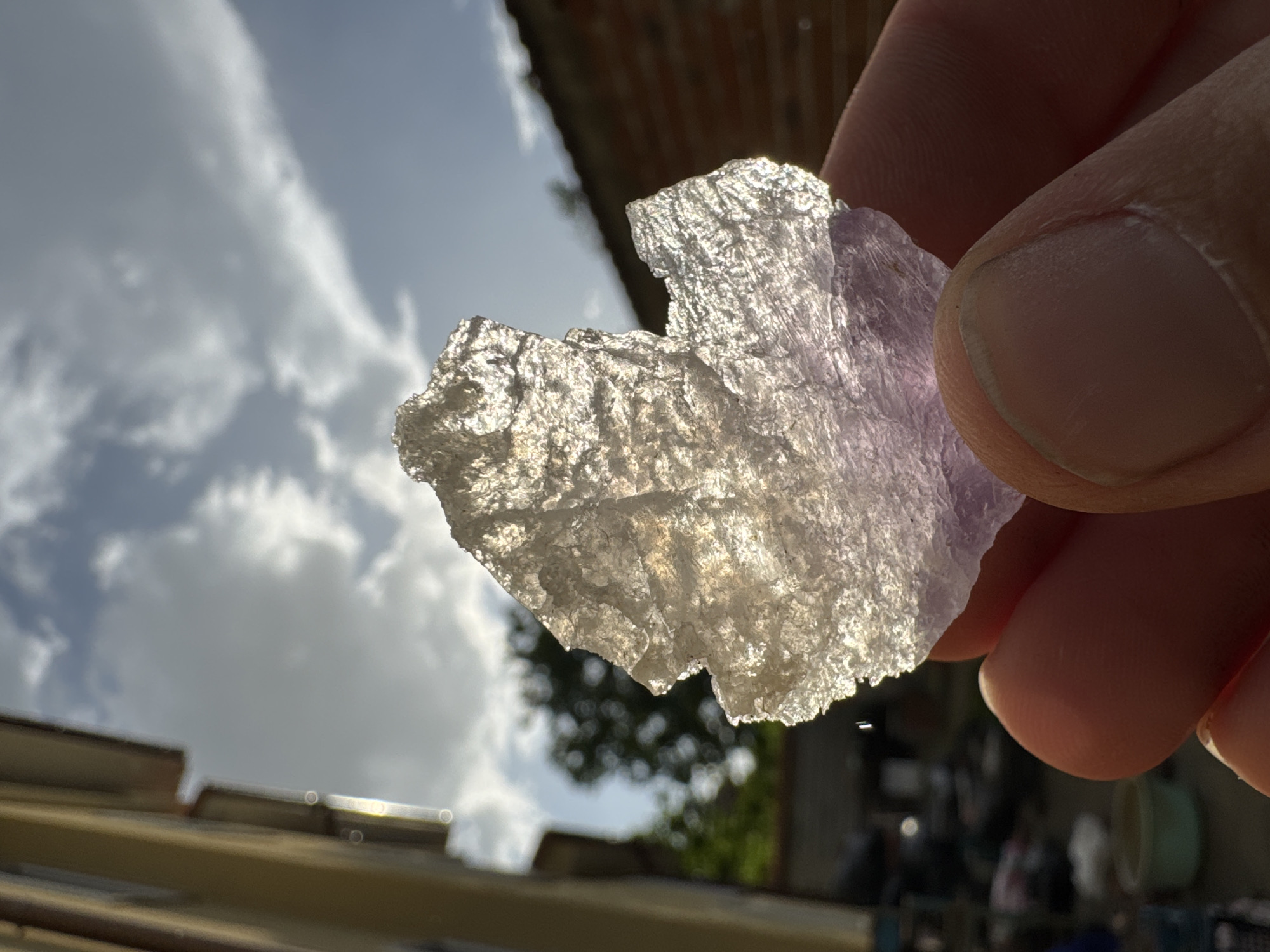 A close-up shot shows a hand holding a translucent, roughly shaped object against a backdrop of a partially cloudy sky. The object, possibly a mineral or crystal fragment, exhibits a slightly purple hue and irregular edges. The hand, with visible fingerprints, is holding the object between the thumb and forefinger. The background consists of a sky with scattered clouds, and glimpses of buildings, trees, and other outdoor elements are visible. The location is not definitively discernible, and no people besides the person holding the object are visible in the frame. The focus is sharp on the object and the hand, with the background appearing slightly blurred.