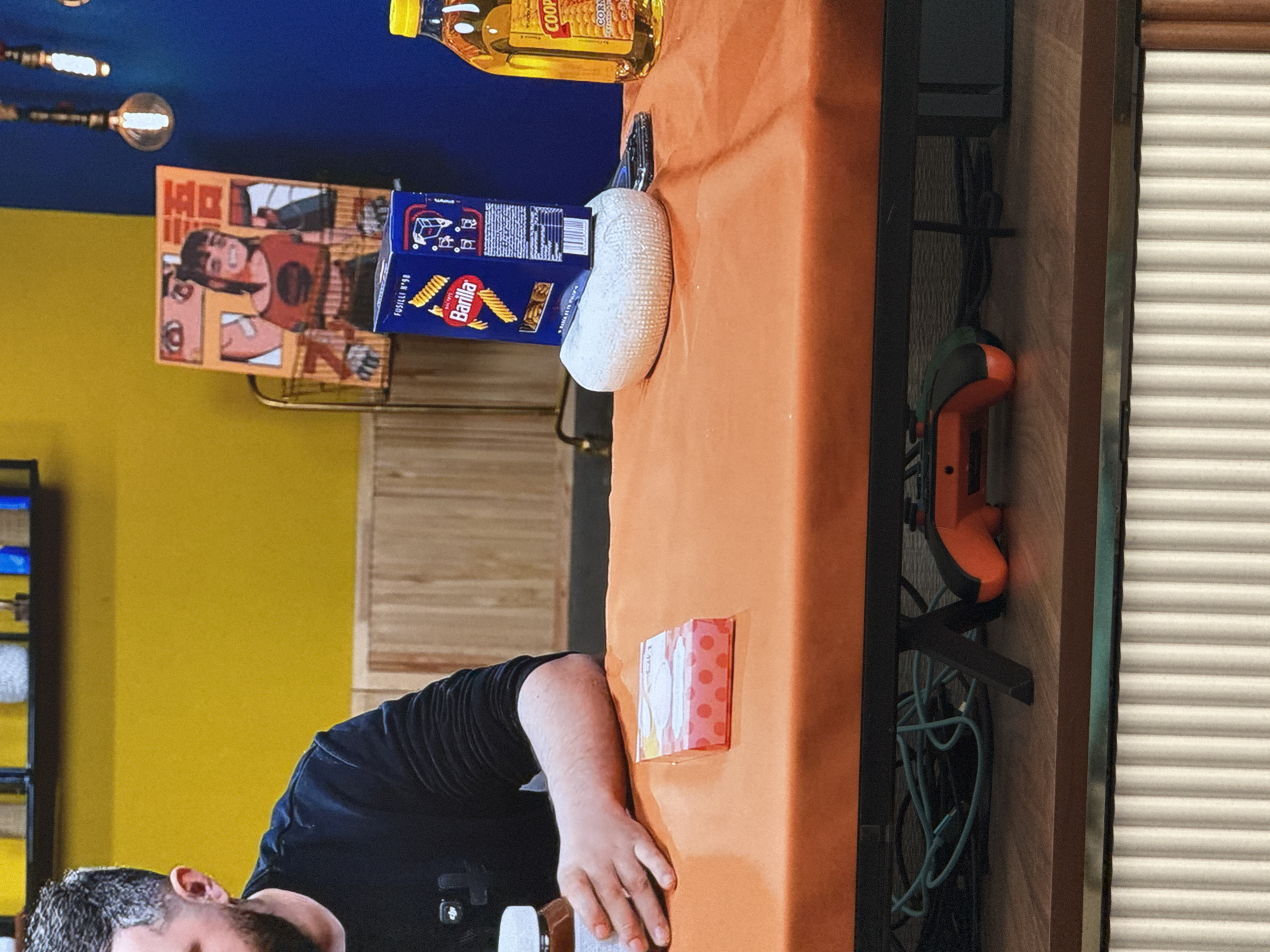 An overhead medium shot captures a person's arm reaching for a jar of what appears to be Nutella on an orange tablecloth. To the right of the jar sits a small, pink box with white polka dots. Further to the right, a blue box of Barilla pasta is displayed on a white, rounded object. Adjacent to the pasta box, a bottle of Cooper's Corn Oil with a yellow cap is visible. In the background, a yellow wall is punctuated by a framed anime-style drawing and a blue wall with exposed lightbulbs. The scene is set indoors, possibly in a studio or kitchen, with a wooden panel wall in the middle ground and a corrugated white wall in the background. A gaming controller rests on the shelf beneath the orange tablecloth.
