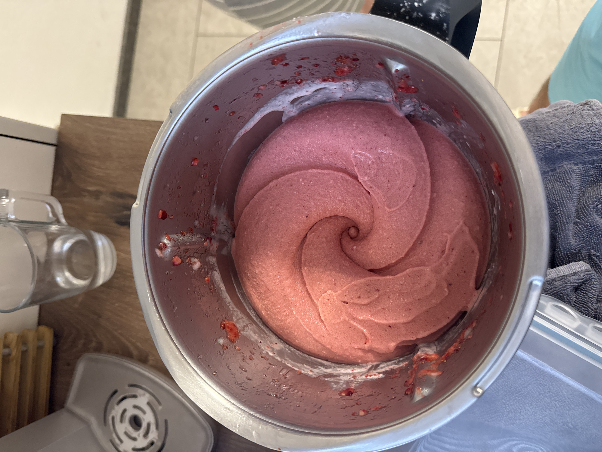 A close-up, overhead shot shows a spiral of pale pink, creamy strawberry ice cream in a silver mixing bowl. The ice cream has formed a perfect swirl, with smaller chunks of strawberries visible around the edges of the bowl. The bowl is sitting on a wooden countertop, with a blurry background that includes a glass of water and part of a kitchen appliance. The lighting is bright, and the overall impression is one of freshness and homemade goodness.