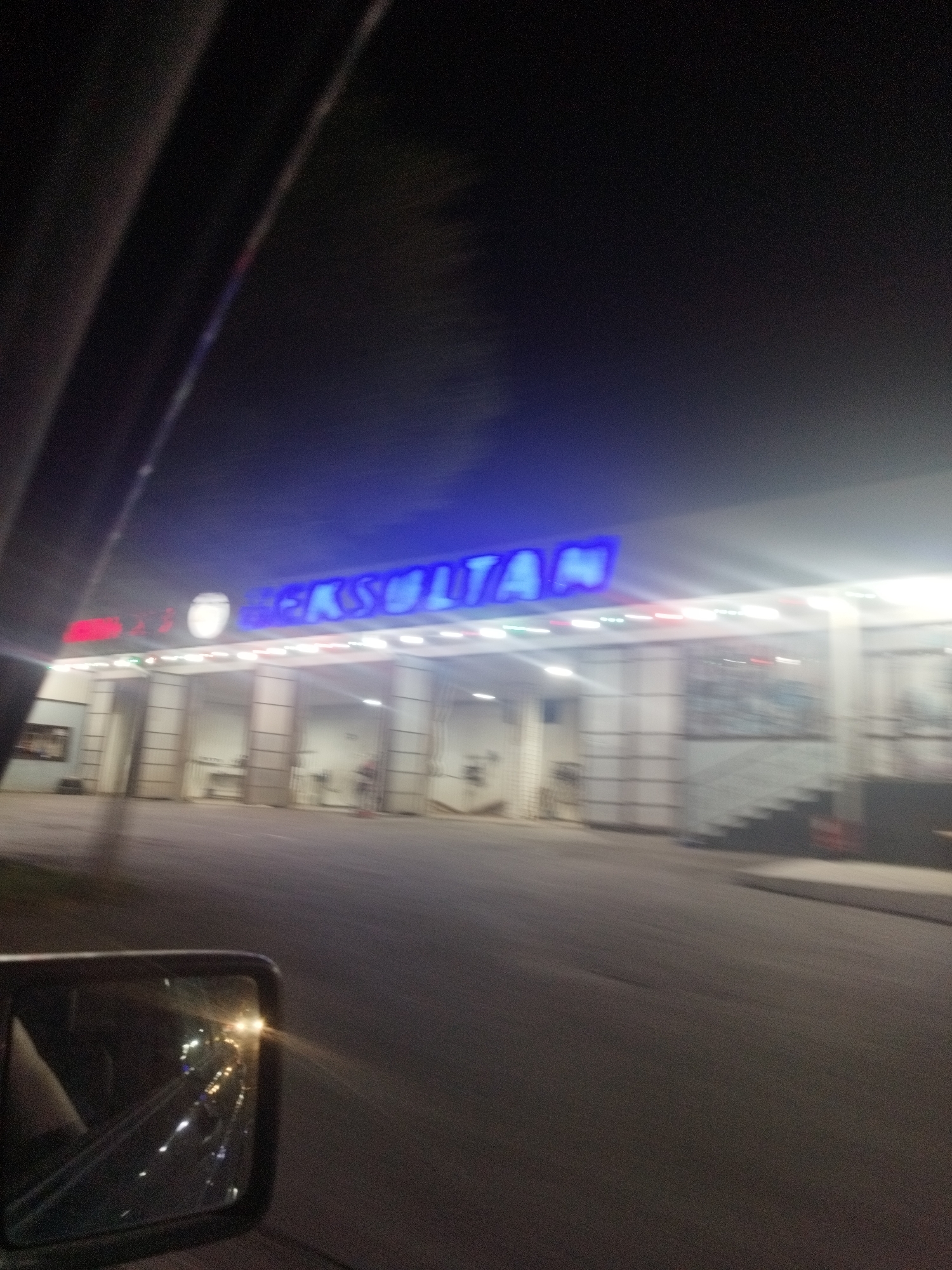 The image, taken at night, shows a building with neon lights in the city of Shymkent, Kazakhstan. The bright blue neon sign above the entrance reads "BEKSULTAN". The building appears to be a commercial establishment with an open-fronted area supported by columns. Lights are visible along the facade. A car's side mirror is in the foreground on the left, reflecting some of the lights from the scene. The image is blurry due to motion.
