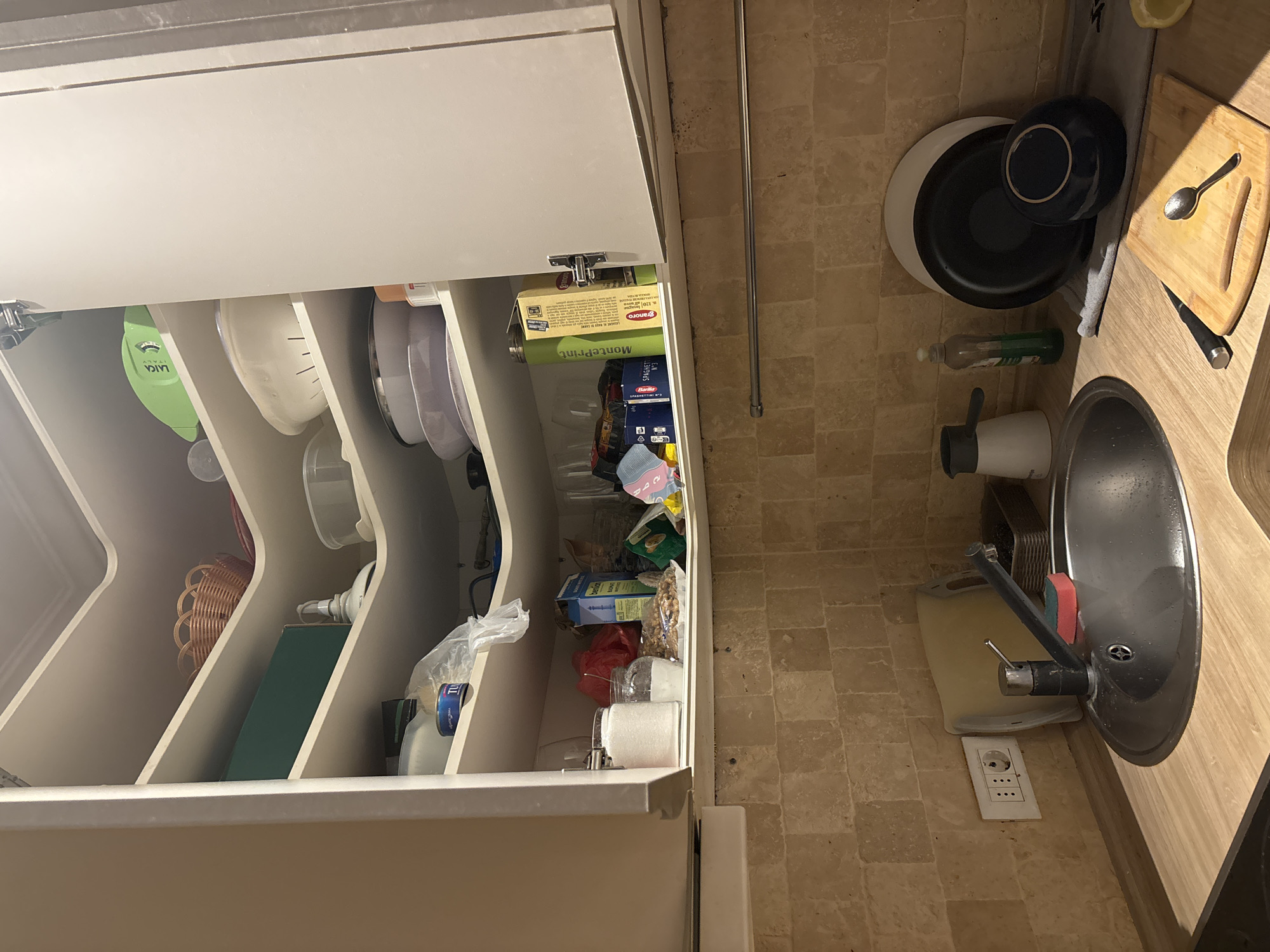 This image captures a kitchen scene, likely in Budva, Montenegro. The viewpoint is from slightly below eye level, looking up into an open corner cabinet filled with various kitchen items. The cabinet shelves are staggered and curved, typical of a corner unit.
On the top shelf, a woven basket is visible, along with a green electronic device labeled "LAICA ITALY". Below that, several white bowls and plastic containers are stacked. Further down, on a lower shelf, are more food containers, bags of groceries, jars of what appears to be sugar, and a bottle of olive oil. A blue box labeled "SPAGHETTI" is also present. The cabinet doors are open, revealing the contents.
The area below the cabinets shows a kitchen countertop. A stainless steel sink with a faucet and a red sponge is prominent. To the left of the sink, a white electrical outlet with multiple sockets is mounted on a beige tiled wall. To the right of the sink, a small chopping board with a knife and a spoon is laid out, with a stack of plates and a dark blue bowl placed behind it. A bottle of dish soap and a white electric kettle are also on the counter. A yellow lemon is visible on the far right edge of the counter. The overall impression is of a functional, slightly cluttered kitchen space.