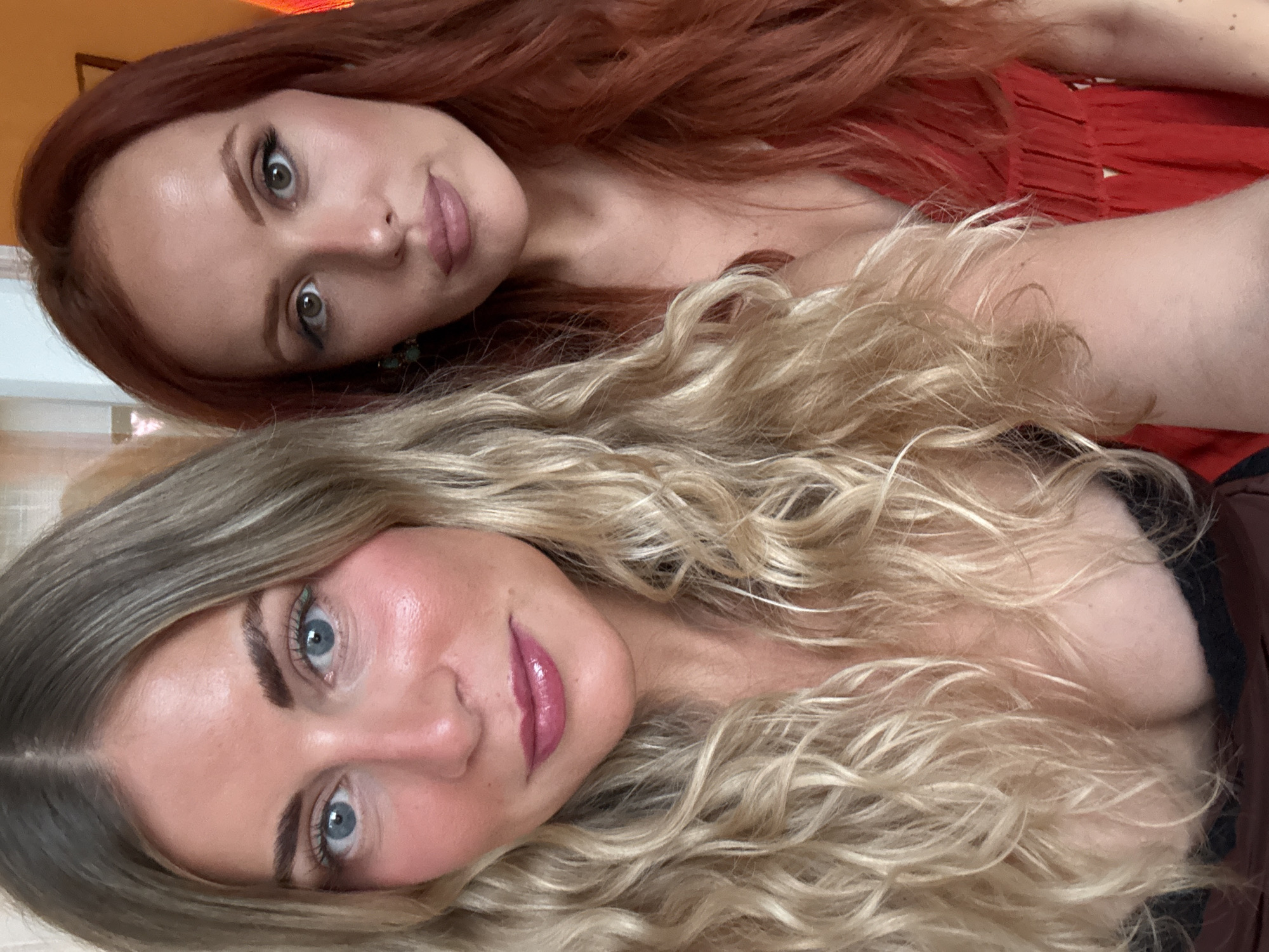 Two women pose closely together for a selfie. The woman at the bottom has blonde, wavy hair, blue eyes, and is wearing pink lipstick. The woman at the top has auburn hair and brown eyes. Both appear to be wearing makeup. The background is indistinct, but there is a glimpse of an orange wall in the upper left corner. The location in Paris is not discernible.