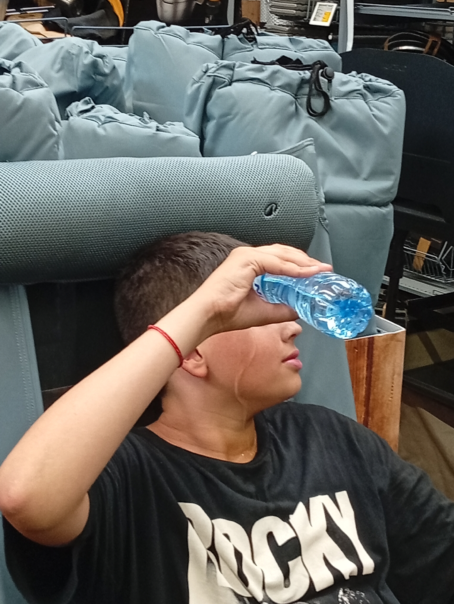 The image shows a young person sitting in a chair, holding a bottle of blue liquid to their forehead. The person is wearing a black t-shirt with "ROCKY" printed in large white letters across the chest. They have a red bracelet on their left wrist. The background is filled with light blue, drawstring bags, stacked on top of each other, suggesting a retail or storage environment. The lighting appears to be artificial, with no clear indication of the time of day. The person's expression is not fully visible but their posture suggests they are attempting to cool down. The scene is likely indoors, possibly in a store or a warehouse in Plovdiv, Bulgaria.