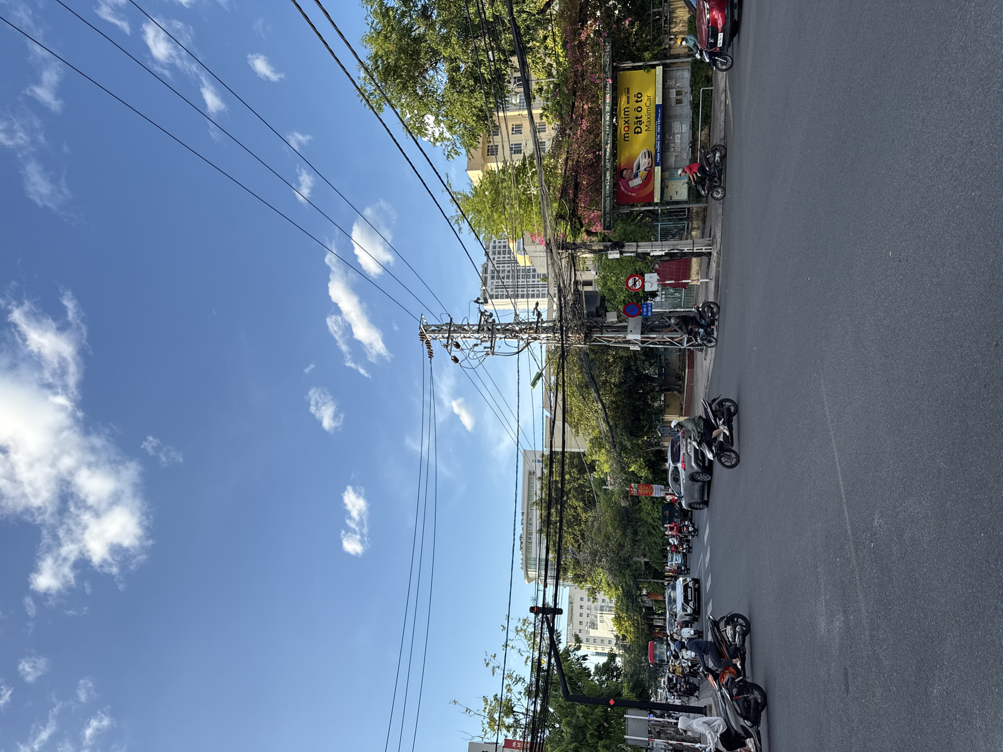 The image depicts a busy intersection in Nha Trang, Vietnam, under a clear blue sky with scattered white clouds. The foreground shows an asphalt road with white lane markings. Several motorcycles and a few cars are navigating the intersection, indicating moderate traffic flow. People are seen riding motorcycles, some wearing helmets.
In the middle ground, trees line the street, and various buildings, including multi-story structures, are visible behind them. A prominent yellow billboard advertising "Maxim" and "Đặt ô tô" (Book a car) is situated on the right side of the road. Electrical poles with numerous tangled wires are a significant feature of the urban landscape. Traffic lights are visible, with one showing red.
In the background, more buildings, including taller modern structures, rise above the trees, suggesting a developed urban environment. The overall scene captures a typical day at a bustling city intersection in Vietnam.