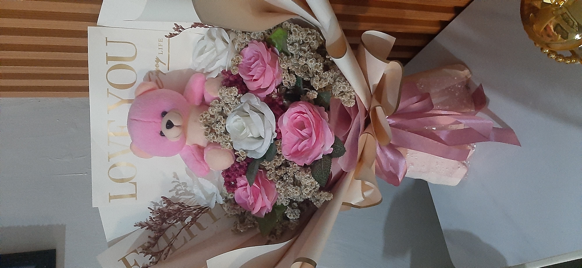 A bouquet of flowers and a stuffed bear are displayed on a table. The bouquet is wrapped in pale pink paper and tied with a pink ribbon. It contains pink and white roses, as well as dried flowers. A small pink stuffed bear sits on top of the bouquet, with a card behind it that reads "LOVE YOU". The bouquet is placed on a white table, with a blurred background of a wall with vertical wooden slats and a hint of a grey wall to the left. There is no indication of the city of Jambi, Indonesia.