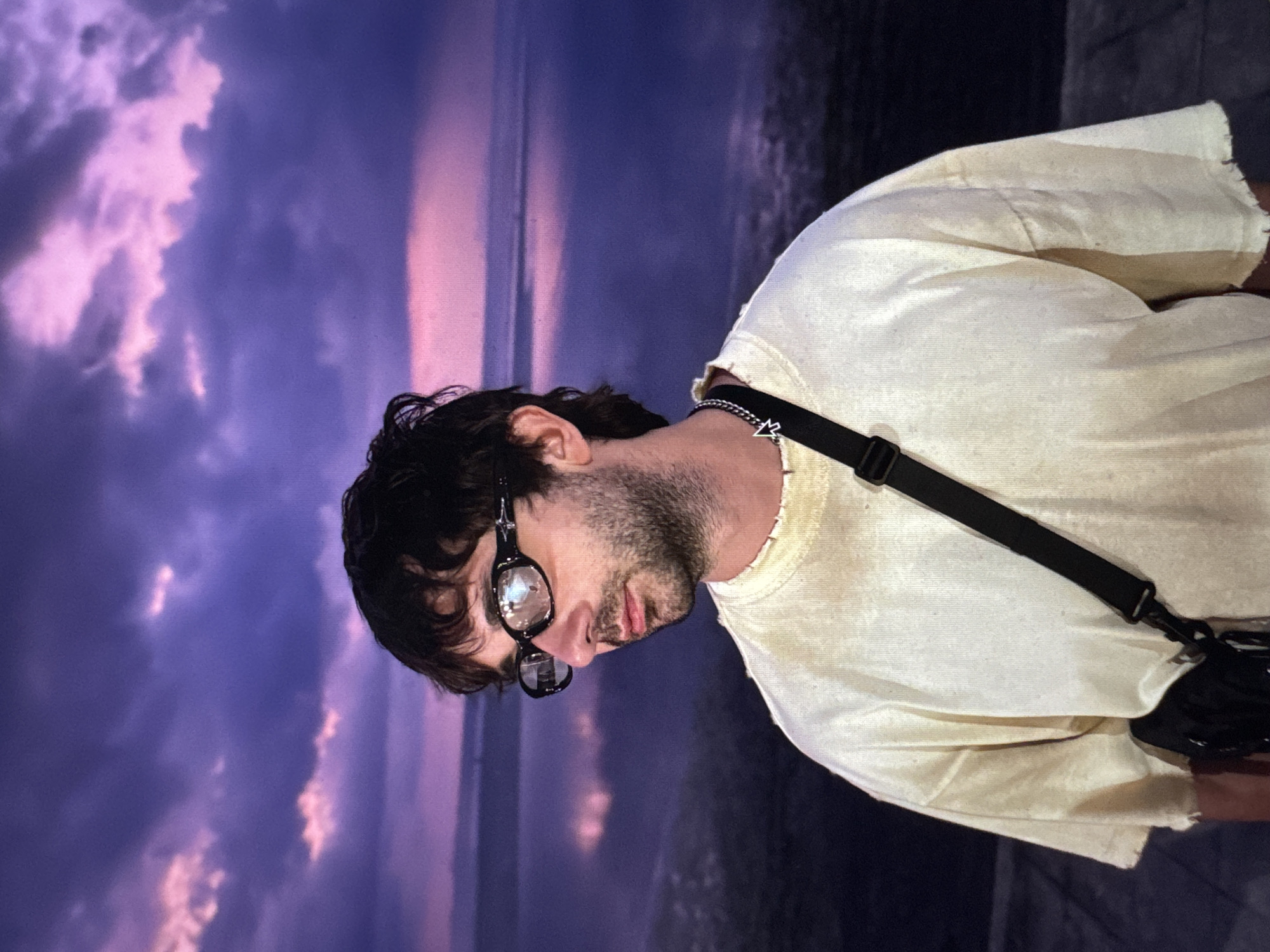A medium shot captures a young man with dark, tousled hair and a beard, wearing glasses and a light-colored t-shirt, against a backdrop of a purple and pink sky reflected in a body of water. The man is looking to his left, his expression neutral. He has a black crossbody bag slung over his shoulder and is wearing a silver chain necklace. The background appears to be a natural landscape, possibly a lake or sea, with a dramatic sky. There is no indication of this scene being in Moscow, Russia.