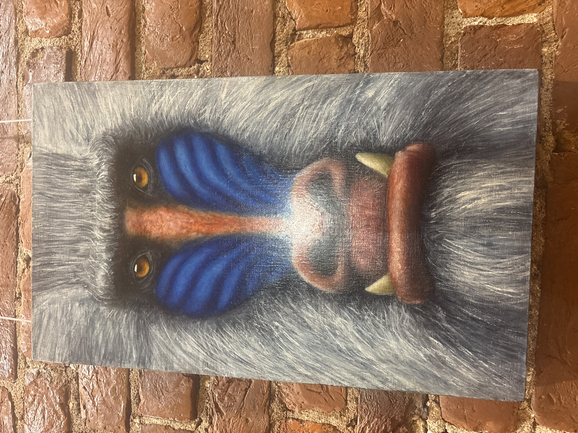 A close-up, vertical oil painting of a baboon's face hangs on a brick wall. The baboon's fur is depicted in shades of grey and white, with prominent blue and orange markings on its face. Its eyes are a striking amber color, and its mouth shows two visible yellow tusks. The painting is illuminated, and the brick wall provides a textured background. The location is described as being in Saint Petersburg, Russia, though the specific context or exhibit is not provided.