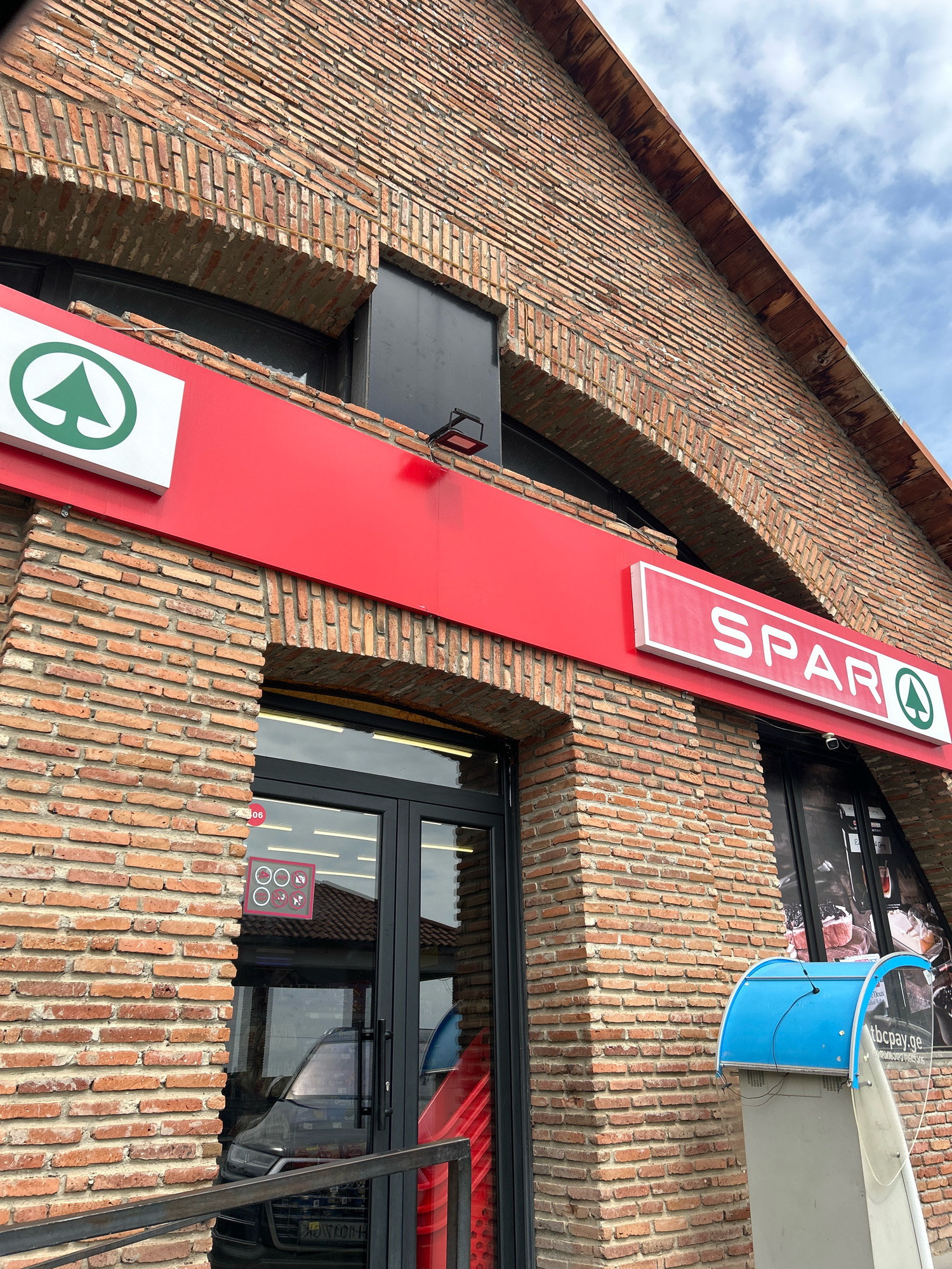 The image shows the exterior of a Spar supermarket in Sighnaghi, Georgia. The building is constructed of red brick with arched windows and wooden eaves. A red sign with the Spar logo is prominently displayed above the entrance. The entrance itself has glass doors, and through them, a black SUV can be seen parked inside. To the right of the entrance, a blue and gray ATM machine stands outside. The sky is cloudy.