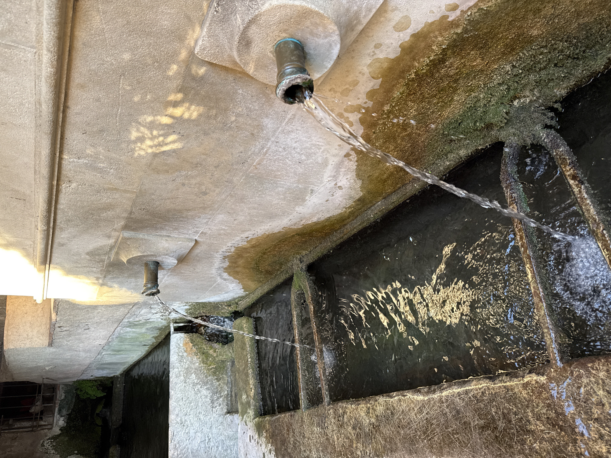 The image shows an outdoor public fountain in Moustiers-Sainte-Marie, France. Two spigots are mounted on a stone wall, with water flowing from them into tiered rectangular basins below. The stone is weathered and has patches of green algae and mineral deposits. The water is clear and reflects light. One basin appears to be overflowing into the next. The overall environment suggests a rustic and historic setting, typical of a Provençal village. There are no people visible in the image.