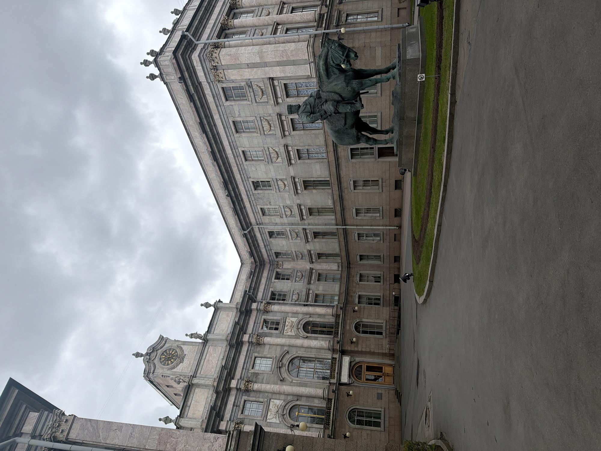 An eye-level shot, originally rotated, shows a grand, multi-storied historic building under an overcast sky in Saint Petersburg, Russia. The impressive structure features neoclassical elements, including numerous windows, prominent columns, ornate cornices, and decorative relief sculptures. The facade appears to be constructed from light-colored, possibly marbled, stone. On the left side of the building, a section rises higher, crowned with a clock tower displaying a golden clock face with Roman numerals.
In front of the building, slightly to the right of the center, stands a large equestrian statue. It depicts a man, possibly a military figure, in a uniform riding a horse. The statue is made of dark bronze.
The foreground consists of a wide paved area, likely a road or public square, with a neatly manicured strip of green grass bordering the building's base. A small sign with a white circle and a red diagonal line (a "no entry" or "no standing" symbol) is visible in the grass. The cloudy sky suggests an overcast daytime, contributing to a muted light across the scene. The image captures a monumental and historically rich urban landscape.
