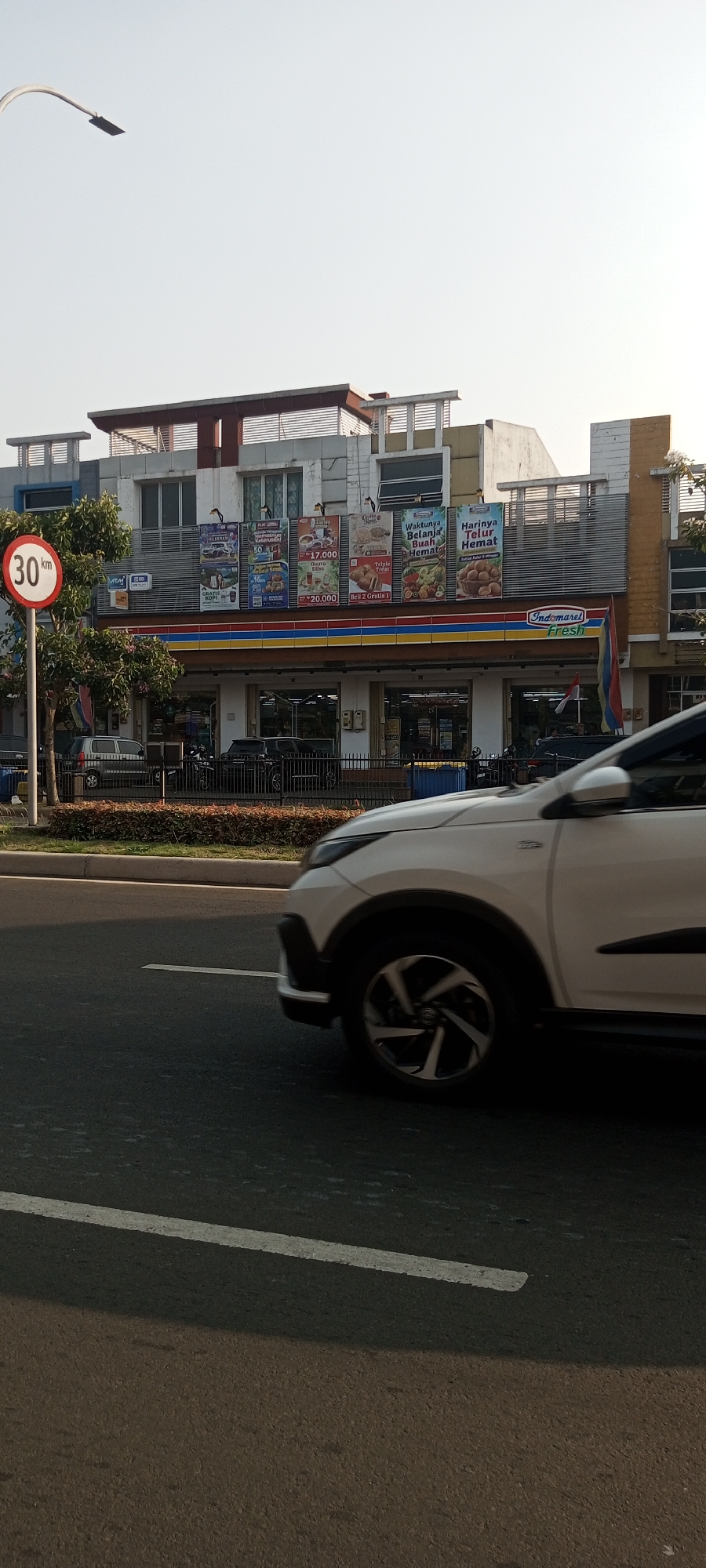 The image shows a street scene in Lambangjaya, Indonesia, on a clear day. In the foreground, a white car is driving on the right side of the road. The car is a modern SUV with alloy wheels. The road has a white line marking the lane. In the midground, there is a sidewalk with a speed limit sign indicating 30 km/h. Behind the sidewalk is a row of buildings, which appear to be shops or businesses. One of the buildings is a brightly lit Indomaret store, a common convenience store chain in Indonesia. Several advertisements are displayed on the front of the Indomaret store, featuring various promotions. There are also some trees and decorative bushes along the sidewalk. In the background, more buildings are visible, some with awnings and balconies. The sky is a light blue, indicating daytime. Several parked vehicles, including cars and motorcycles, are visible in front of the shops. There are no people clearly visible in the image.