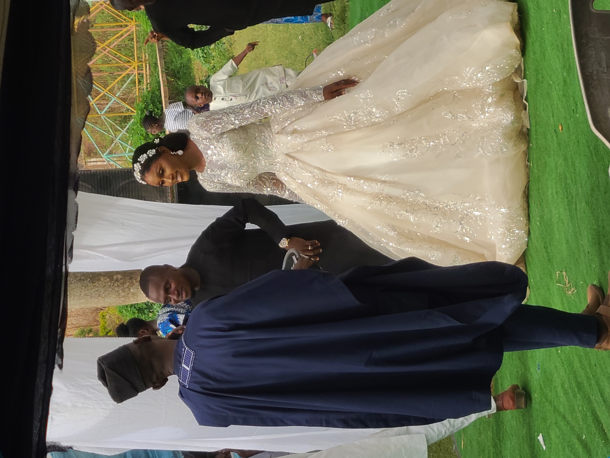 The image shows a wedding scene in an outdoor setting, likely in Jos, Nigeria, given the attire and environment. A bride in a long-sleeved, cream-colored wedding gown with silver embellishments is the central figure. She wears a headpiece and is smiling. Several other individuals are present. A man in a dark blue traditional outfit with a dark-colored cap stands prominently in the foreground. Another man in a black suit is visible behind the bride. In the background, people wearing casual clothing are visible. The environment appears to be a grassy area with a white backdrop possibly made of fabric and an overhead canopy or structure. The quality of the image indicates it was likely taken with a mobile phone.