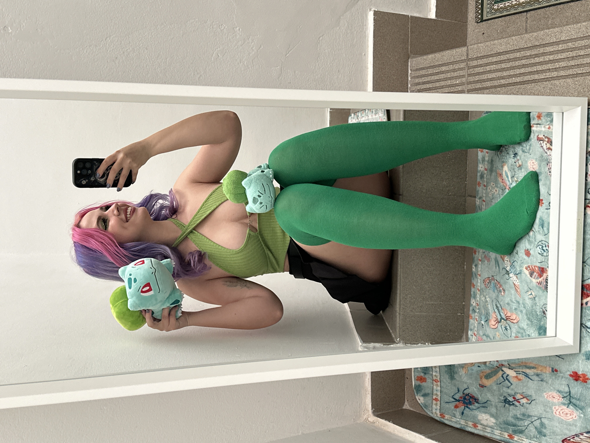 A woman with pink and purple hair is holding a light blue and green stuffed animal that resembles Bulbasaur. She is wearing a green tank top and green thigh-high socks, and she is taking a photo of herself in a mirror. The mirror is propped up against a wall, and behind the mirror is a set of tiled steps and a rug with insect patterns. The location is within a building in City of Zagreb, Croatia.