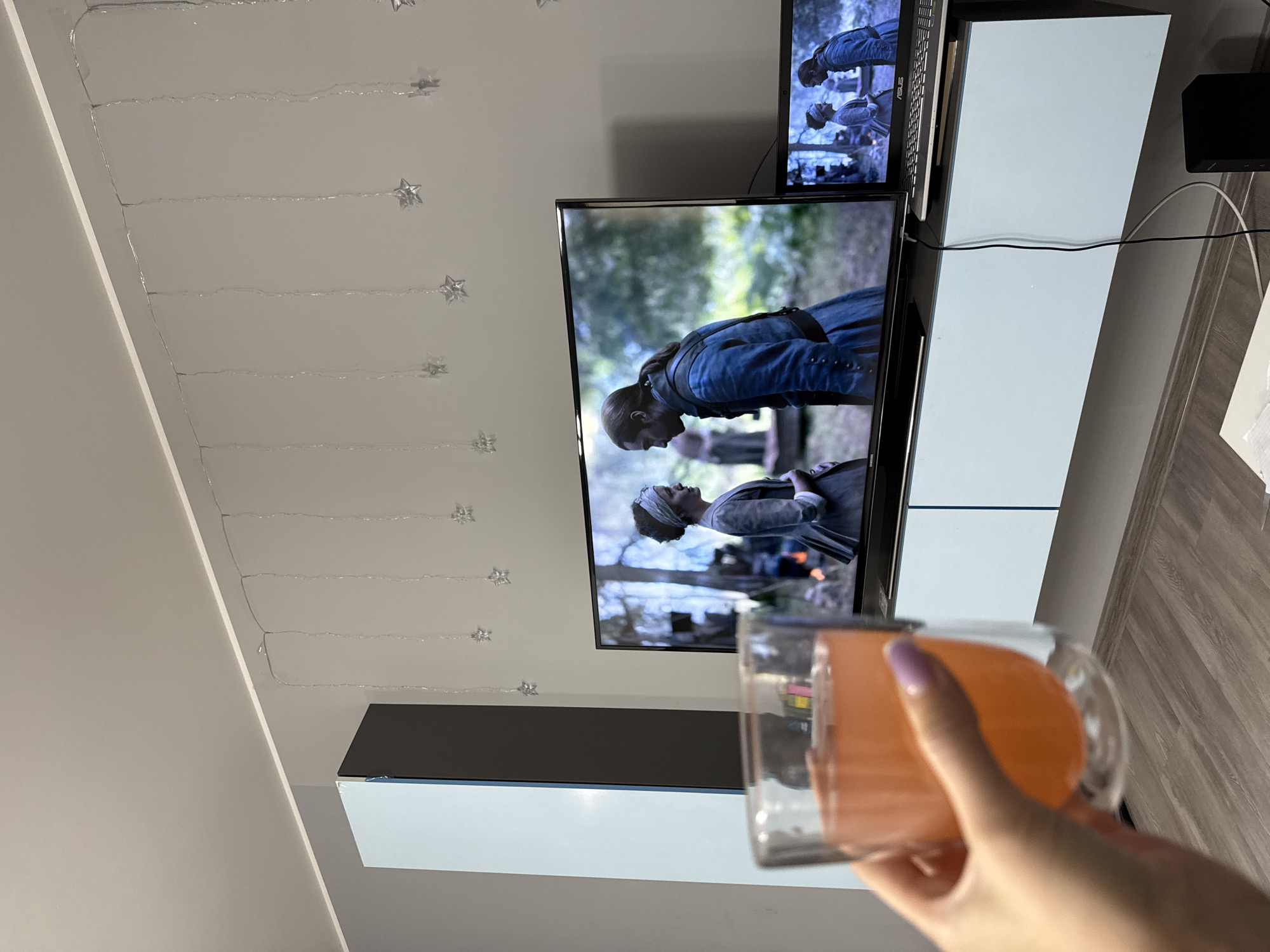 The image shows a living room scene. A hand holding a glass of orange-colored liquid is in the foreground, to the left. In the midground, a large flat-screen television displays a scene from a historical drama, featuring two actors in period costumes. To the right of the television, a laptop is also displaying the same scene. The television and laptop are placed on a light blue media console against a light gray wall. A string of small, star-shaped lights is visible on the wall above the television. A tall, dark gray and light blue cabinet is partially visible on the left side of the image. The floor is made of light brown laminate. The image does not provide any specific location information about Łódź, Poland.