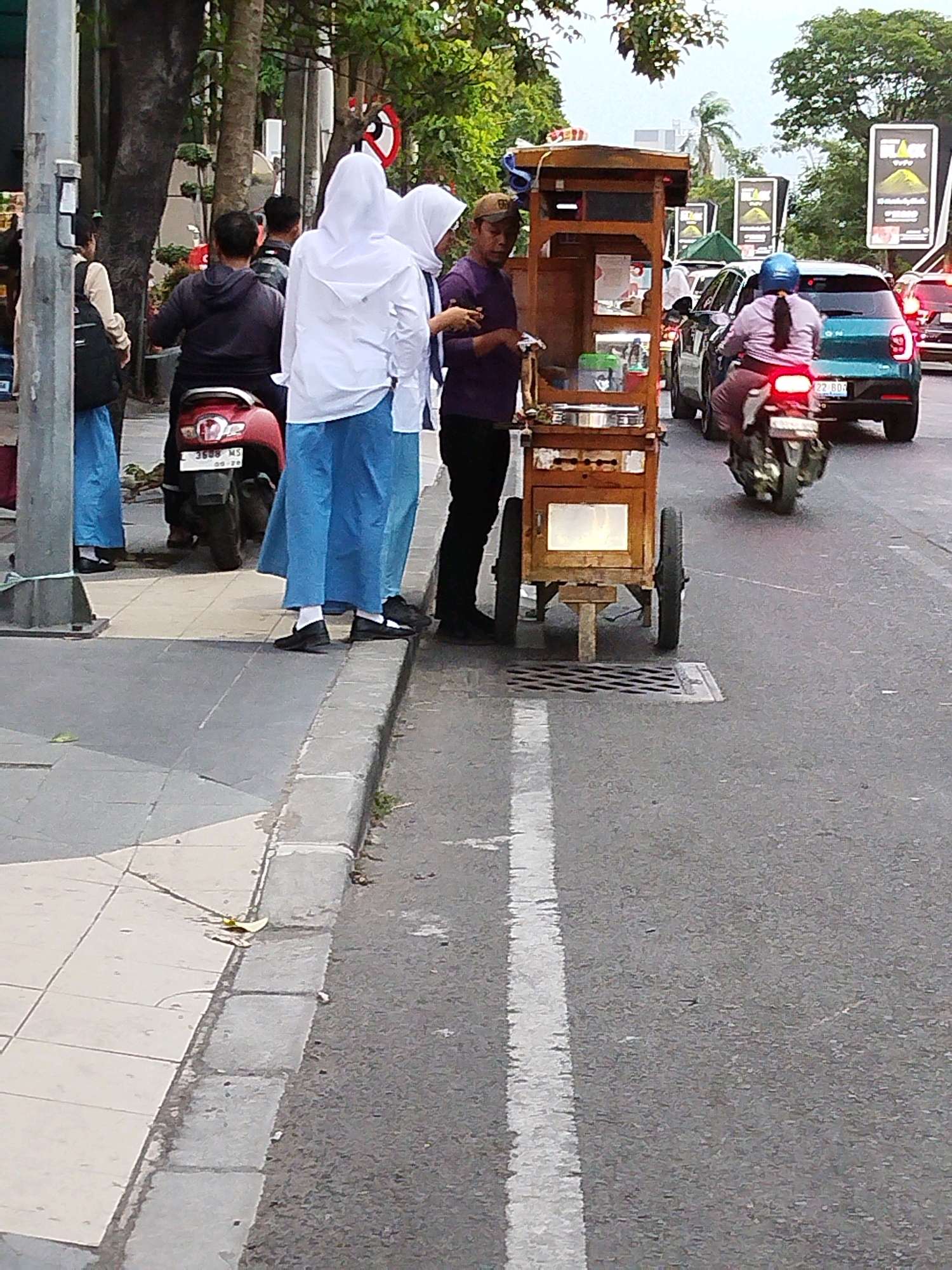 In Surabaya, Indonesia, a street scene unfolds with a vendor's cart in the foreground. The wooden cart, a mobile food stall, has a tall wooden display unit with two transparent compartments on the upper section, likely for ingredients or finished products. Below these compartments is a light-colored surface, possibly a warming tray or preparation area, with several items in containers. The cart stands on two large wheels at the rear and two smaller wheels at the front, with a support leg extending downwards. A man wearing a purple shirt and a brown cap stands behind the cart, facing left, holding a small white item. To the left of the cart, two individuals in white hijabs and blue skirts stand on a tiled sidewalk. One is facing the cart, while the other is looking to the left. A red motorcycle is parked behind them, facing left. Further down the street, a woman wearing a blue hijab and a pink top rides a motorcycle, heading away from the camera. She is in the right lane of the road, passing by a teal car. The road has a white line marking the lane. In the background, trees and buildings are visible, and several billboards are mounted along the street. The overall impression is a busy street scene in an urban environment.