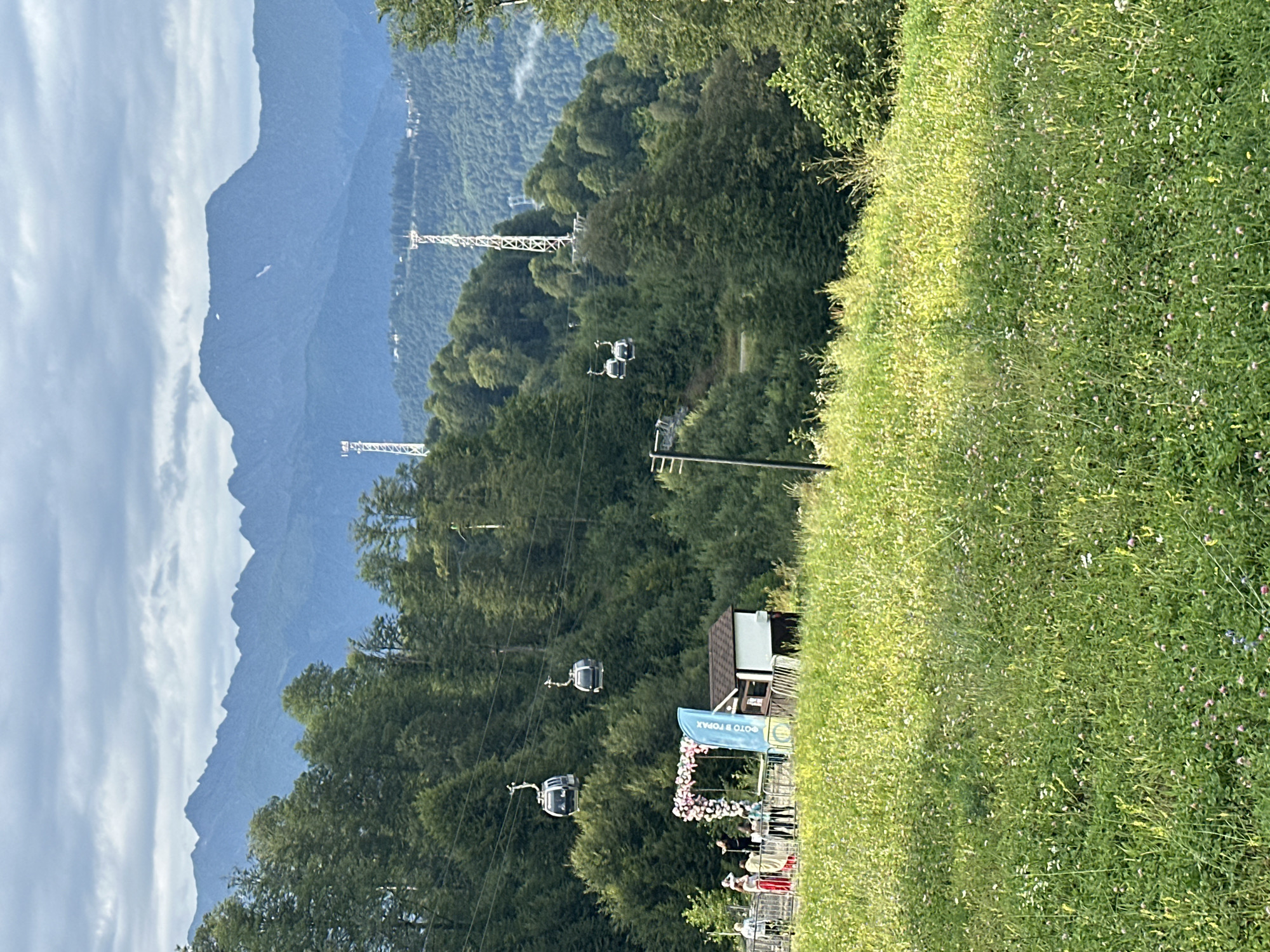 The image depicts a scenic view in Estosadok, Russia, featuring the Krasnaya Polyana ski resort area. In the foreground, a lush, green meadow dotted with small wildflowers slopes upwards. In the midground, a group of people are gathered near a decorative floral archway, possibly for a photo opportunity. A small building, resembling a ticket booth or information center, is situated next to the arch. Several cable cars are visible ascending a forested mountainside. The background is dominated by layers of tree-covered mountains, culminating in a rugged, mist-shrouded mountain range under a cloudy sky. Tall white communication towers stand on some of the higher slopes. The overall atmosphere is tranquil and picturesque, indicative of a mountain resort in the summer or late spring.