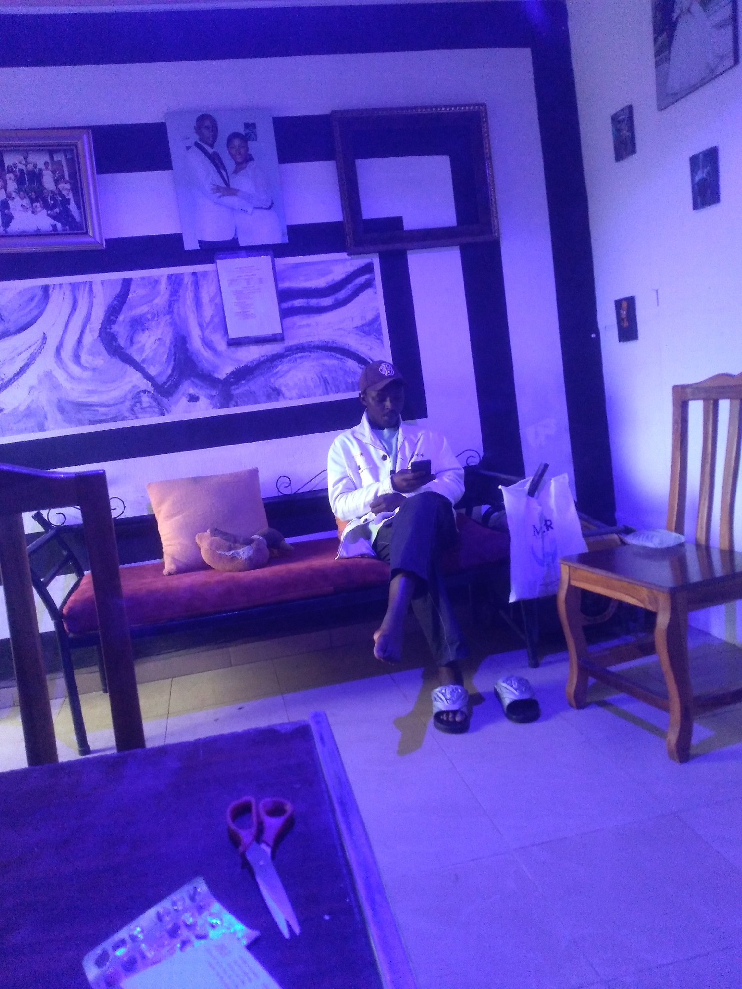 An indoor scene features a man sitting on a long, red-cushioned bench under distinctive blue-purple lighting. He wears a white jacket, dark trousers, and a cap, and is engaged with his mobile phone. His sandals are placed on the tiled floor in front of the bench. Next to him on the bench are a beige cushion and a small, light brown pastry-like item. A white plastic bag with the letters "MR" written on it rests on the bench to his right.
The walls are decorated with framed pictures and artwork. Behind the man, a section of the wall features bold black and white vertical stripes, while another part has a large abstract painting with swirling blue and purple patterns. Framed images visible include a group photo on the left, a portrait of a couple in the center, and several smaller, darker frames on the white wall to the right. A document or sign in a small frame is also mounted on the wall below the couple's portrait. A wooden chair stands to the right of the bench. In the foreground, a wooden table is partially visible, holding a pair of red-handled scissors and what appears to be a blister pack of pills or small items. The artificial lighting creates a moody, cool-toned atmosphere.