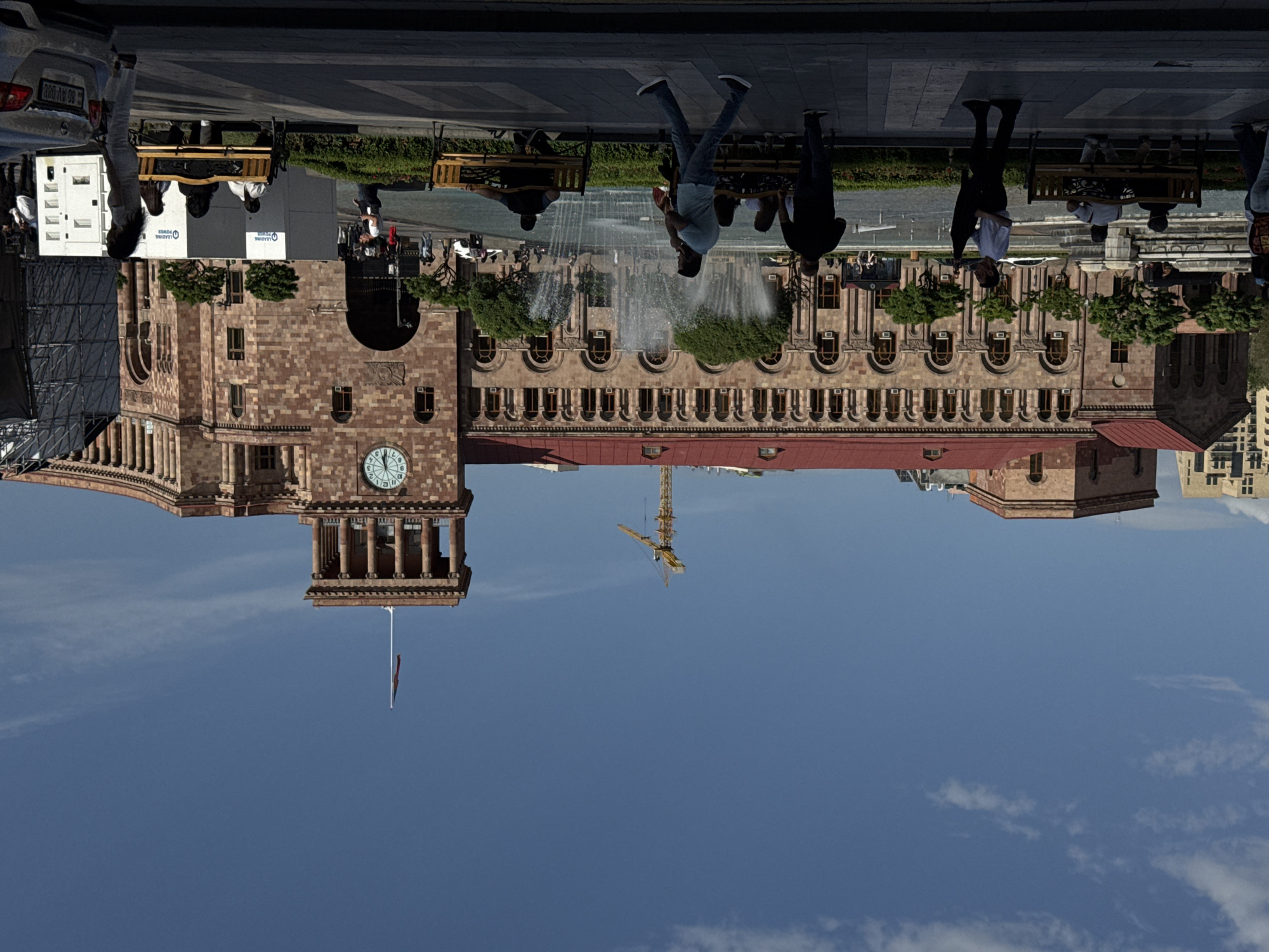 The image depicts Republic Square in Yerevan, Armenia. The scene is taken from an unusual angle, which is why it is necessary to rotate the image to view it accurately. At the top, people are walking and sitting on benches along the plaza. A large fountain is in operation, and the buildings of Republic Square are visible in the background. A construction crane is visible, and the sky is clear with some clouds. A clock tower and an Armenian flag are prominently featured.