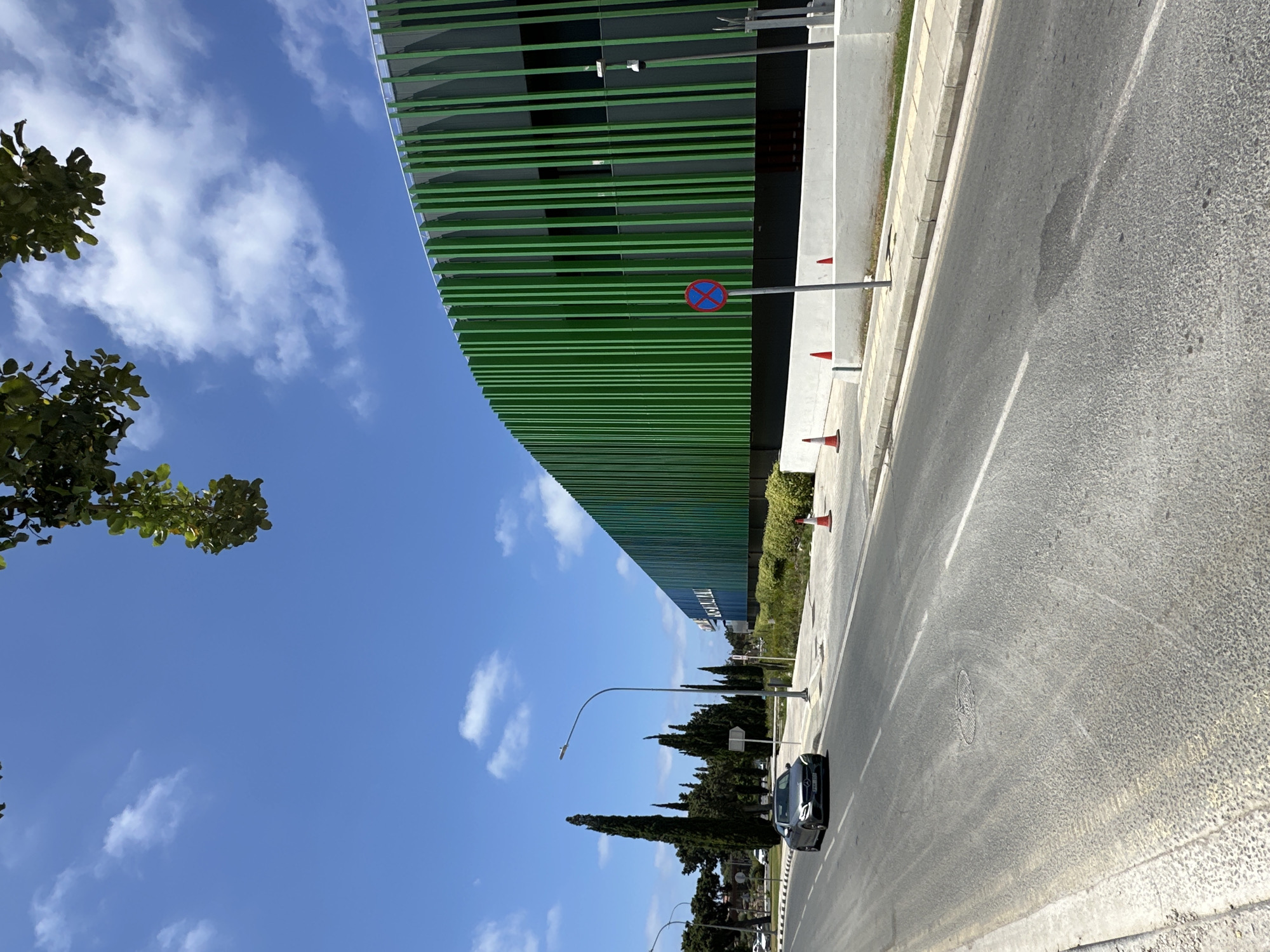 The image shows a modern building with a green slatted facade in Paphos Municipality, Cyprus. A grey Mercedes-Benz car is driving on the asphalt road in front of the building. The sky is blue with scattered white clouds. Several tall cypress trees are visible on the left side of the road. There are traffic cones and a "no stopping" sign near the building. The building appears to be a commercial or public facility. The scene suggests a sunny day in an urban or suburban environment.