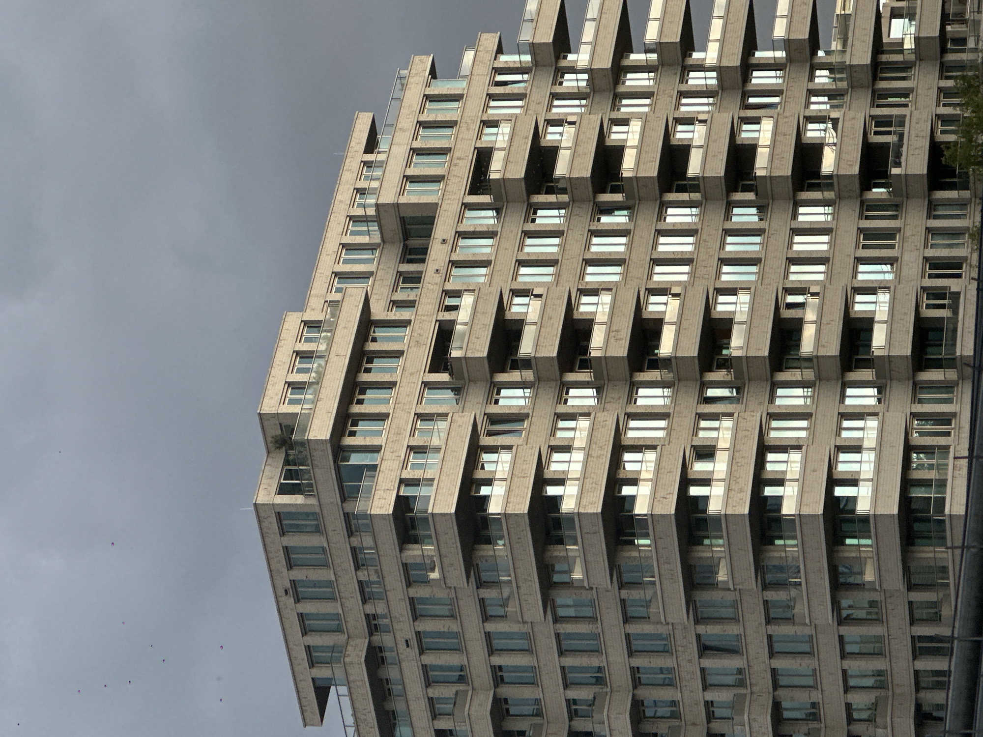 The image shows a tall building, likely a residential complex, in Amsterdam, Netherlands. The building is light gray and constructed with a unique design: each floor juts out slightly, creating a staggered, vertical pattern that defines the facade. The windows are rectangular and reflect the sky, which is overcast and gray. The building occupies the right side of the frame, while the left side is dominated by the gray sky. The angle of the shot is from below, looking upward. A few specks, possibly birds, are visible in the sky. Vegetation, likely trees, is visible on the far right edge of the building. The building's architecture is the most prominent feature.
