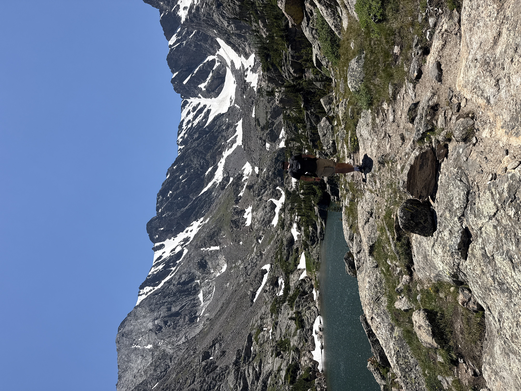 An adult male wearing a black t-shirt, tan shorts, and a black backpack walks on a rocky trail toward a body of water. The trail is surrounded by large rocks and patches of green grass. In the background, a large body of dark blue water is visible, with steep, rocky mountains rising behind it. The mountains are partially covered in snow. The sky above is clear and blue. The scene appears to be in a natural, mountainous area, possibly Rocky Mountain National Park, near Allenspark, Colorado, United States.