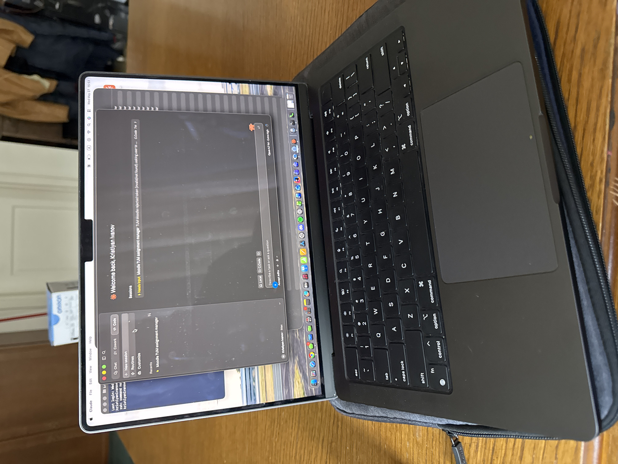 A laptop is open on a wooden table. The laptop screen displays a chat application, potentially a development environment, with the name "Kristiyan Ivanov" visible. The laptop's keyboard and trackpad are visible. A case surrounds the laptop. A box of OMRON products is partially visible on the left side of the image. The date "Wed May 27 15:53" is displayed on the laptop screen. The location is undetermined, but the chat messages suggest "TUM" and the last login suggests "kristiyanivanov". The city of Sofia, Bulgaria is not visually evident in the image.