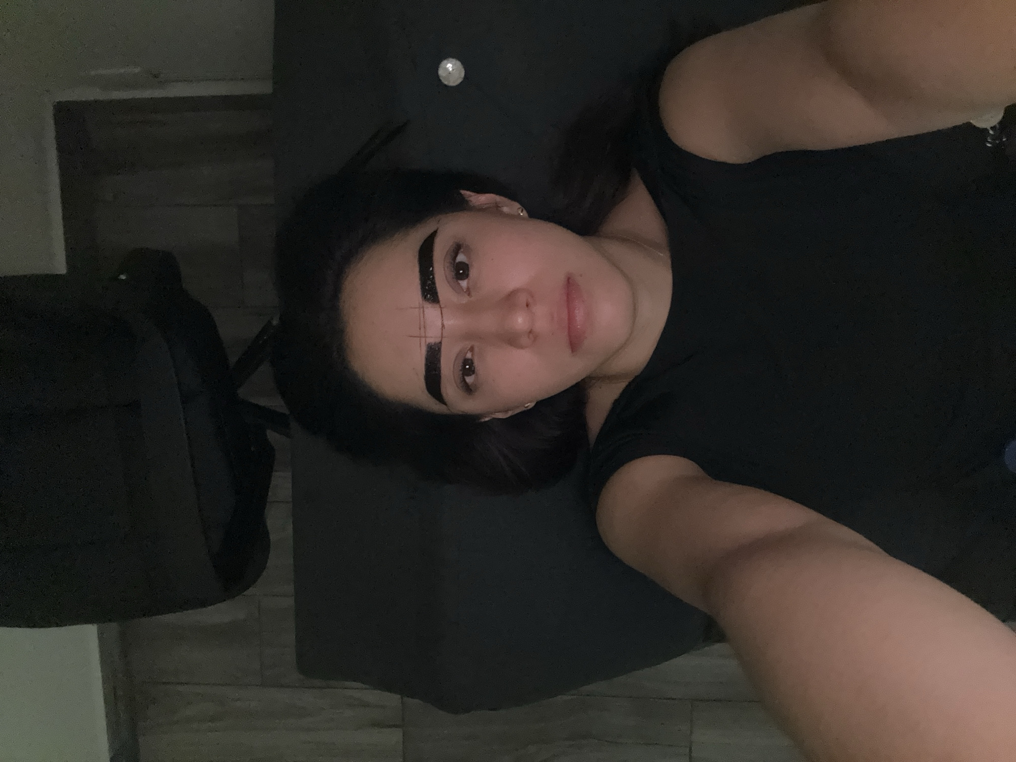 A young adult female lies on her back on a dark surface, possibly a mat or cushion. She wears a black t-shirt. Dark tape is applied to her eyebrows, with markings drawn on the tape. A small, shiny object is visible near her head on the dark surface. Part of a dark-colored chair and a light-colored tiled floor are visible in the background. The location, Puebla, Mexico, cannot be determined from the image.
