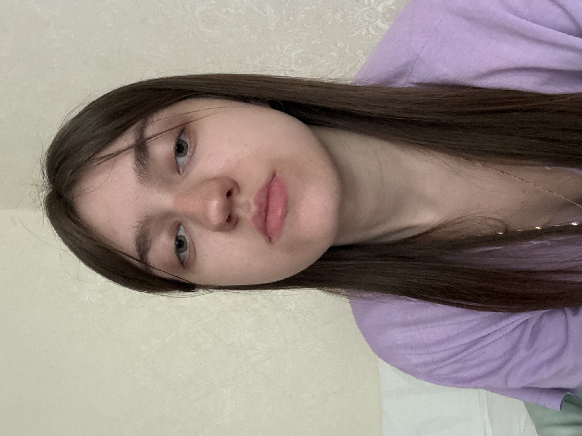 A close-up, slightly rotated, indoor portrait of a young woman with long, straight, brown hair and fair skin. She has light green eyes, full lips, and a small nose. She is wearing a lilac-colored top. The background consists of a light beige wall with a subtle floral pattern. No city or precise location can be deduced from the information.