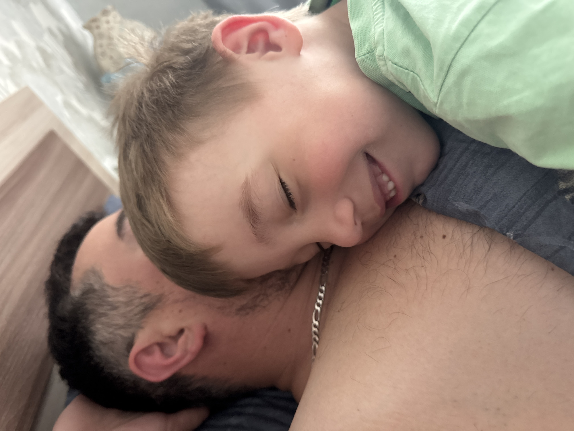 A close-up, indoor shot shows a child, wearing a light green top, lying on a man’s chest. The man has short, dark hair and a trimmed beard. A silver chain is visible around his neck. The background appears to be a bedroom, with a bedside table and textured wallpaper. The image does not offer enough detail to definitively confirm the location as the city of Chaikovsky, Russia.