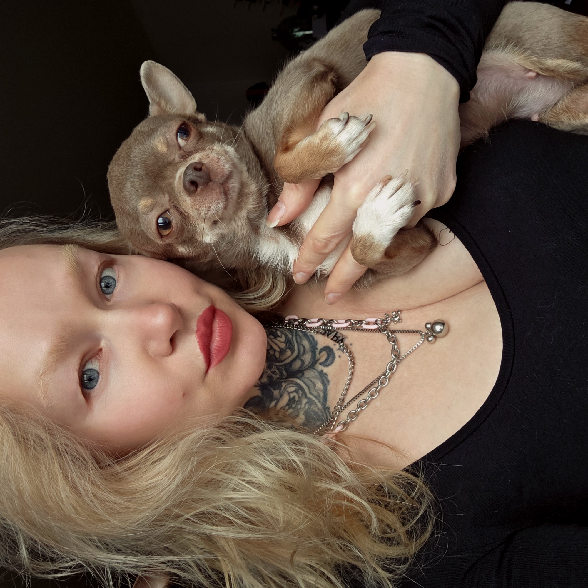 The image presents a close-up, tilted portrait of a blonde woman with blue eyes and bright red lipstick, embracing a small, light brown dog. The woman has fair skin and an intricate black tattoo visible on her chest and collarbone, which includes stylized text that appears to read "BYBA". She wears several layered necklaces, one featuring a pink chain element, another with a heart-shaped charm, and a third with a spherical pendant. She is dressed in a black top.
The dog, likely a Chihuahua, has light brown (fawn) fur with white markings, particularly on its paws. It has a dark nose and dark, expressive eyes, looking directly at the camera with a curious or alert expression. The woman is holding the dog close to her, with the dog's front paws resting on her arm and its belly visible against her body. The intimate pose suggests an affectionate interaction between the woman and her pet. The setting appears to be indoors with soft lighting. No specific outdoor location cues are visible.