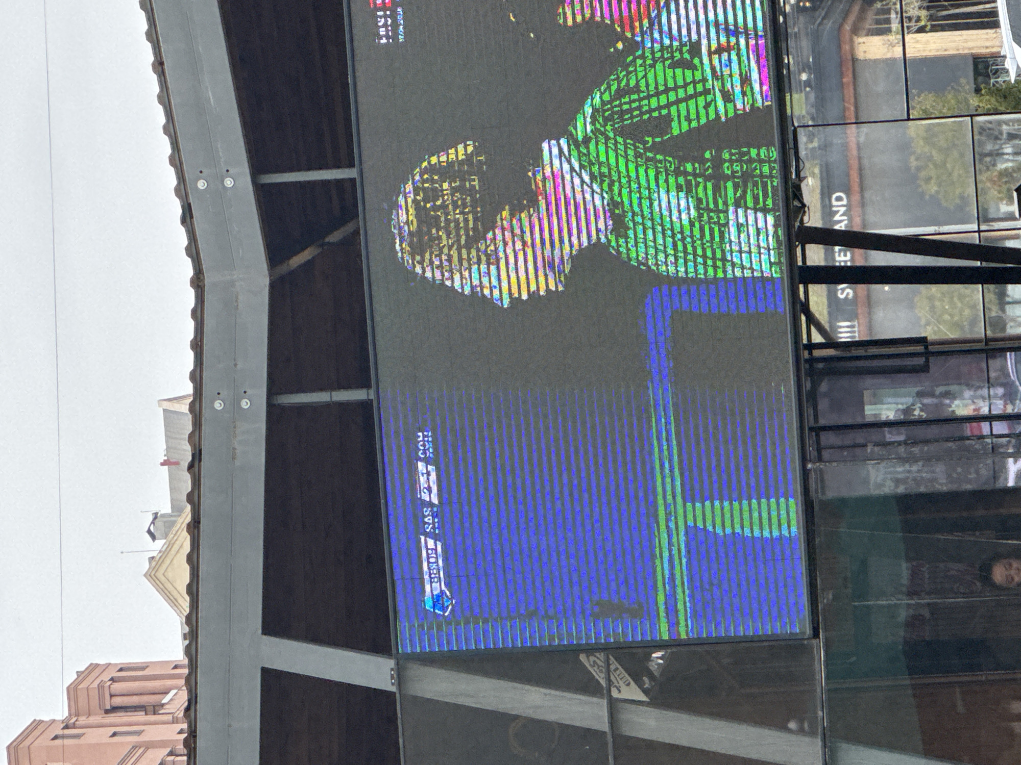 Captured at an angle, the image showcases a large outdoor LED screen displaying a distorted image, likely a video feed, with noticeable vertical banding. The colors are primarily blue, green, and yellow. In the lower left corner, a portion of a building is visible with a pink-beige facade, while the sky appears overcast. Metal structural elements frame the left side of the screen. Reflections on glass surfaces near the screen reveal elements of the surrounding urban environment, including a sign with the words "S EET AND" and a person's face. The image displays an SAS vs FROBER 2-1 COM score. While not definitively discernible, the presence of "SAS" potentially indicates a sports-related broadcast. The presence of Armenian text and the city name being mentioned means it can be presumed this may be the Yerevan city in Armenia. The reflection displays “Sweet and” indicating the presence of shops.