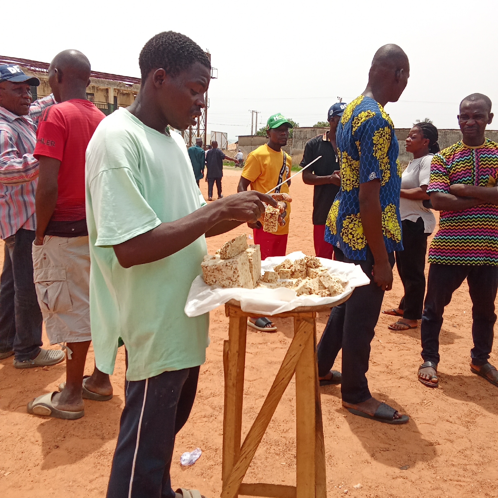 An outdoor scene in Akwanga, Nigeria, shows a group of people gathered on reddish-brown dirt ground under a clear, bright sky, suggesting daytime. In the foreground, a man in a light green t-shirt and dark blue pants stands beside a small wooden stand covered with a white cloth. He holds a piece of what appears to be groundnut brittle or a similar nut-based snack, with more blocks and smaller pieces of the snack arranged on the stand for sale.
Around him, several other individuals are present. To his immediate left, a man in a red t-shirt with "HLER" visible on it stands with his back mostly to the camera, beside another man in a striped shirt and a blue cap bearing faint letters like "DF" or "DE." In the mid-ground, slightly to the right, a man in a yellow t-shirt and a green cap (with "HO" visible) holds a long, thin stick. Behind him and to his right, a man in a blue and yellow patterned shirt faces away from the camera, conversing with a woman in a white t-shirt and black pants. On the far right, another man in a brightly colored, patterned shirt with zig-zag designs stands with his arms crossed.
The setting appears informal, possibly a market or a roadside gathering. Simple buildings are visible in the background, along with some utility structures. Many individuals are wearing sandals. A crumpled white plastic bottle lies on the ground near the wooden stand. The overall atmosphere seems relaxed, with people engaged in interaction and observation.
