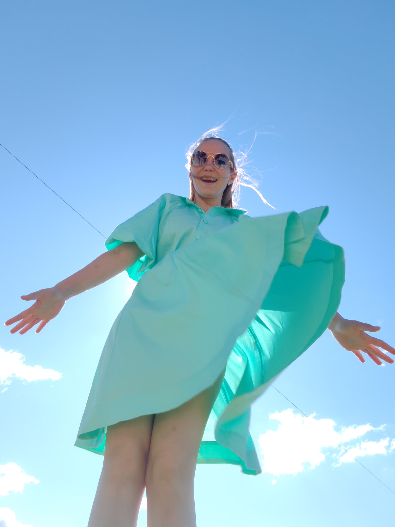 An extreme low-angle, medium shot captures a young woman outdoors under a bright blue sky with scattered white clouds. The sun is positioned behind her, creating a lens flare effect and backlighting her hair, which appears windblown. She is wearing round, gold-rimmed sunglasses and a light teal, short-sleeved dress with a collar and buttons. Her arms are outstretched to her sides, palms facing upwards, and her mouth is open in what appears to be a joyful expression. The hem of her dress is billowing outwards due to the wind, revealing her legs. A thin, dark wire runs horizontally across the upper portion of the frame. The image is a candid shot, likely taken in an open, outdoor location. The specific location in the city of Чухлэм, Russia, is not identifiable from the visual information provided.