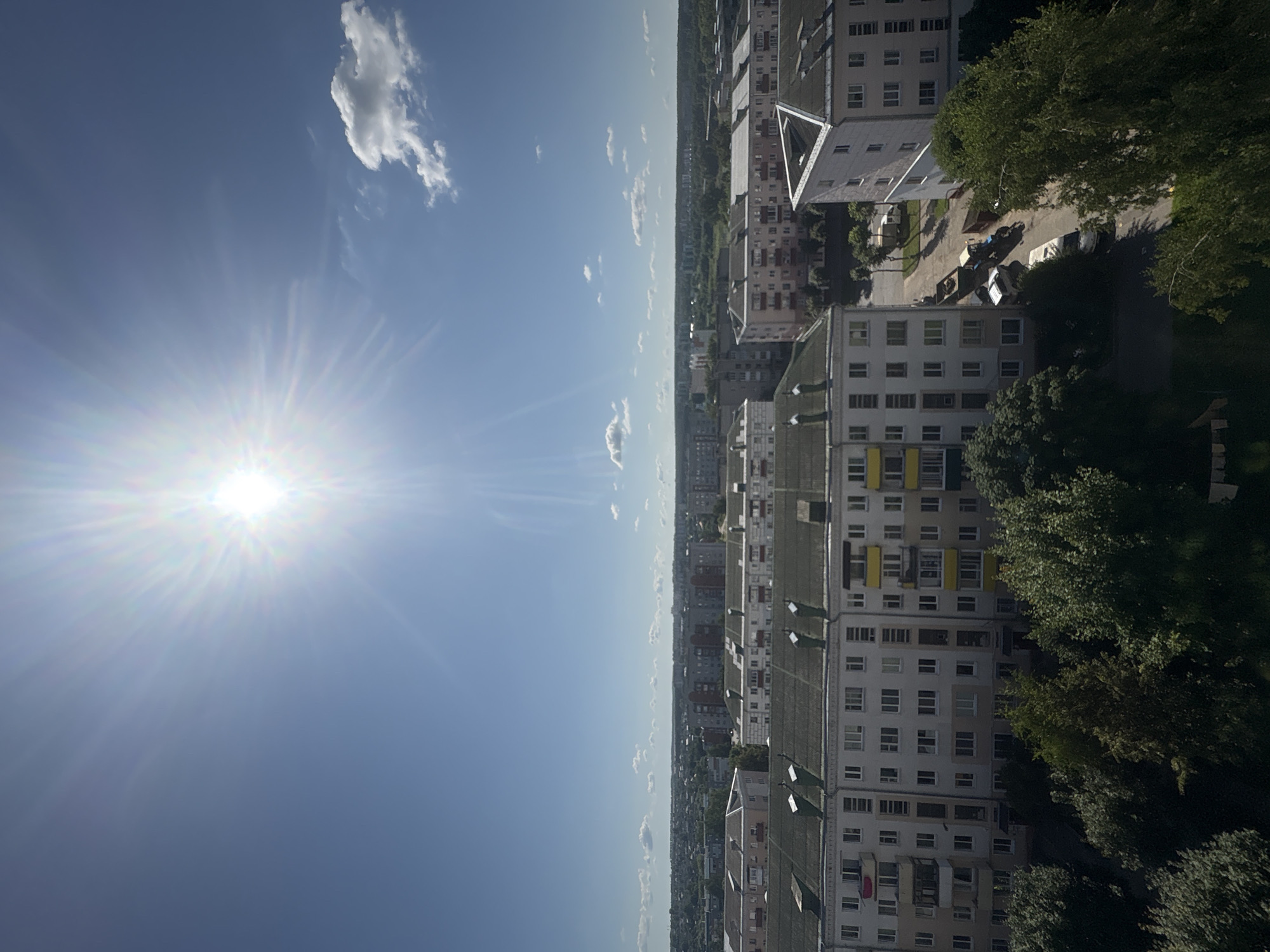The image is a high-angle shot of a city on a sunny day, likely taken from a high-rise building. The sky is a clear blue with a bright sun, which is the main light source and creates a lens flare effect. There are a few scattered white clouds.
In the foreground, there are several apartment buildings with a consistent architectural style, featuring light-colored facades, multiple floors, and balconies. Some of the buildings have distinctive peaked roofs, while others are flat-topped. The rooftops are a mix of grey and white. Below the buildings, there are mature trees with lush green foliage.
The middle ground shows more apartment buildings and a street with parked vehicles, including a blue tractor with a trailer, a white van, and another vehicle. The street appears to be in a residential area.
The background consists of a sprawling cityscape with more buildings of varying heights and colors, extending to the horizon. The landscape beyond the city is dominated by green trees, suggesting a park or forested area.
While the image is clearly of an urban environment, specific identifying features like street signs or distinct landmarks are not visible enough to pinpoint the exact location. However, based on the architectural style and the general appearance of the cityscape, it is consistent with residential areas found in cities like Orsha, Belarus. There are no people visible in the frame. The overall scene depicts a typical day in a European city, with everyday urban infrastructure and elements of nature interspersed.