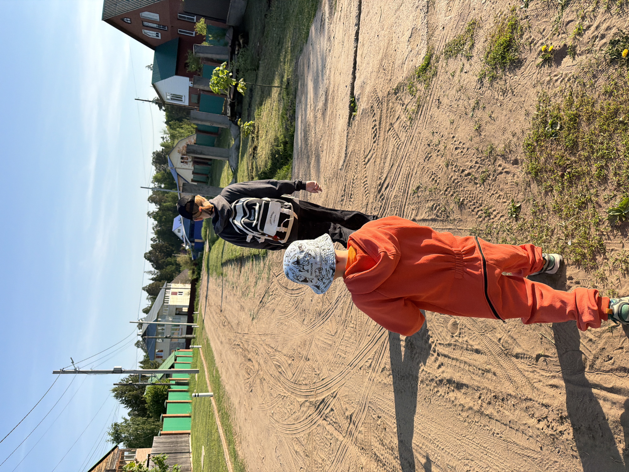 Two individuals are captured in an outdoor setting, likely in a residential area of Chaikovsky, Russia, as suggested by the architectural style of the houses and the utility poles. The foreground features a child in an orange jumpsuit and a patterned bucket hat, while an adult in a black hooded sweatshirt, a baseball cap, and a striped backpack stands behind the child. Both are on a dirt path marked with tire tracks. The environment includes grass patches, and houses with green accents are visible in the background. The sky is clear, indicating a bright, sunny day.