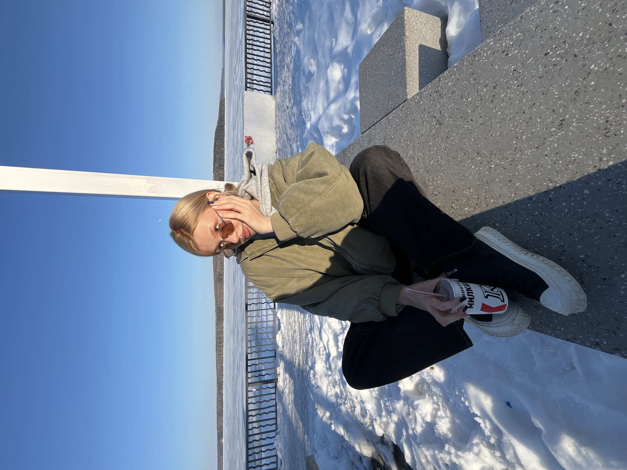 A fair-skinned woman with blonde hair, wearing sunglasses, an olive green jacket, a gray hoodie, and black pants, is sitting on a concrete ledge in Chaikovsky, Russia. She is holding a cup with a straw in her hands. The background includes a snowy landscape, a body of water (likely the Kama River), and a clear blue sky. A black metal railing is visible. A concrete structure sits behind the woman.