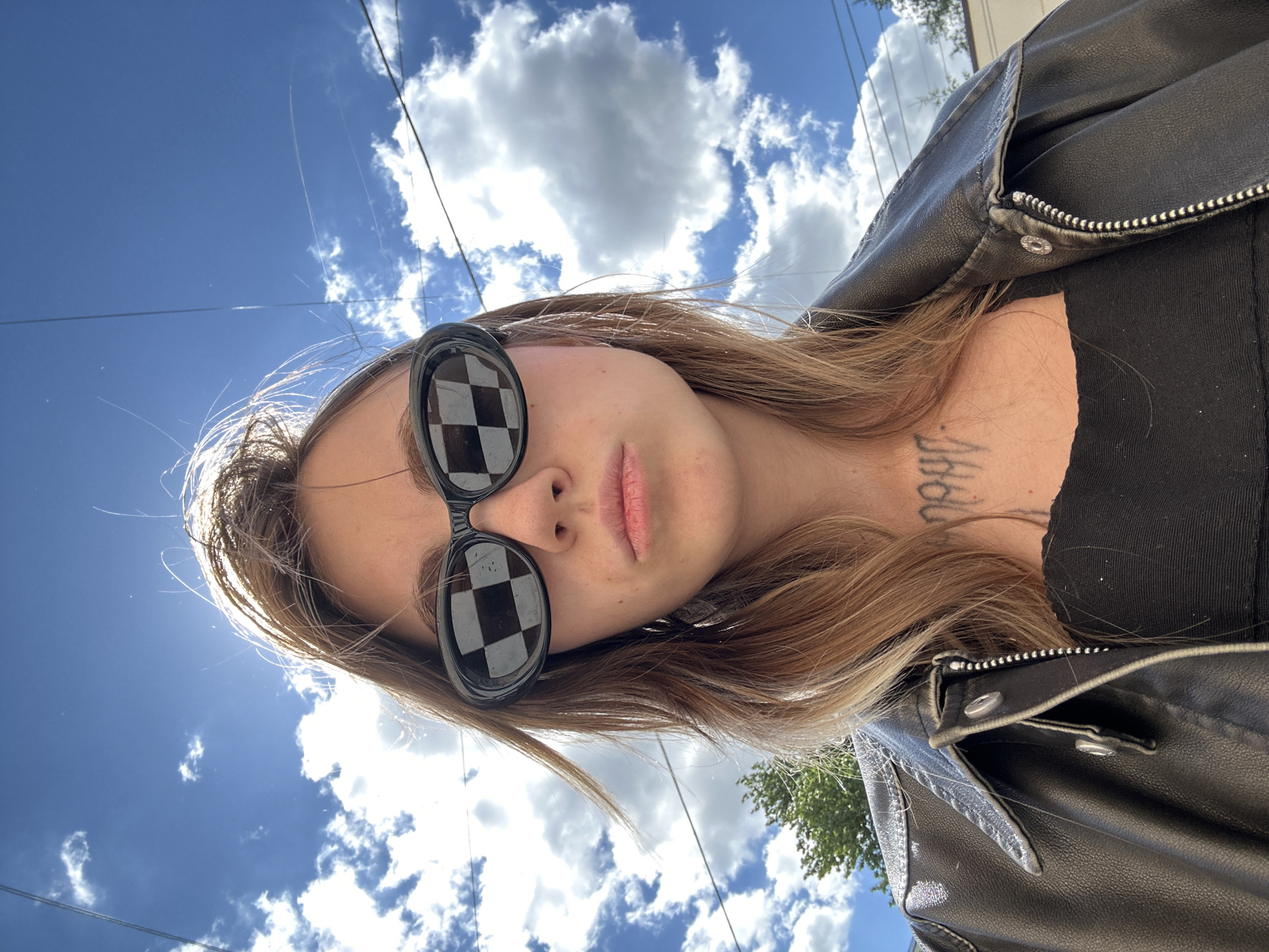 A young woman with long, light brown hair wears black sunglasses with a checkerboard pattern lens and a black leather jacket over a black top. She has a tattoo on her neck. The background is a bright blue sky with white clouds. The image appears to be taken outdoors, possibly during the day.