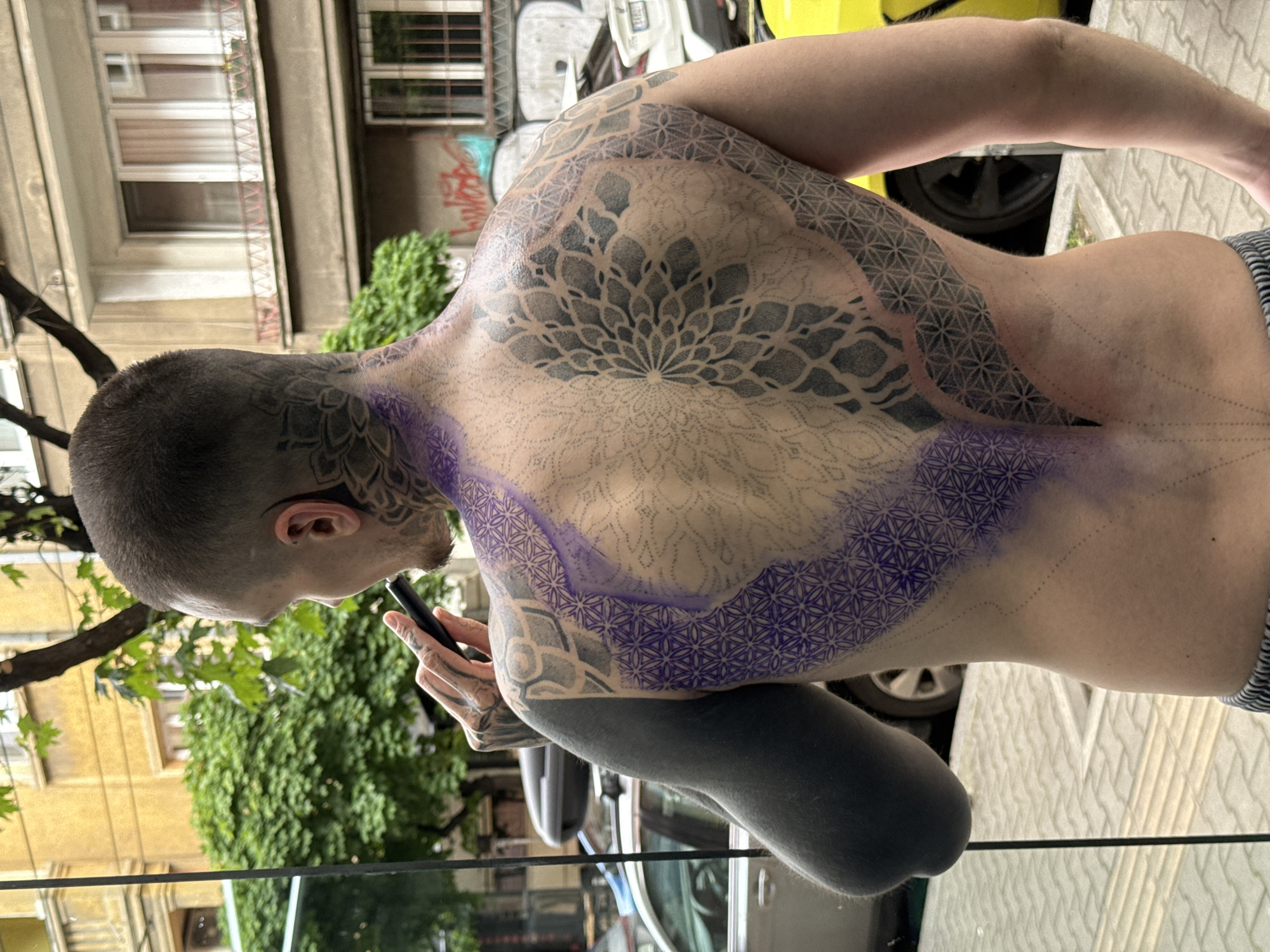 A man with extensive tattoos on his head, back, and arms is standing outside a building in Sofia, Bulgaria, possibly looking at his phone. The tattoos on his back are intricate geometric patterns, with some areas marked in purple, suggesting they are in progress or recently stenciled. He has a full arm tattoo in dark color on his left arm. The background includes a building with graffiti and windows, trees, parked cars and the pavement.