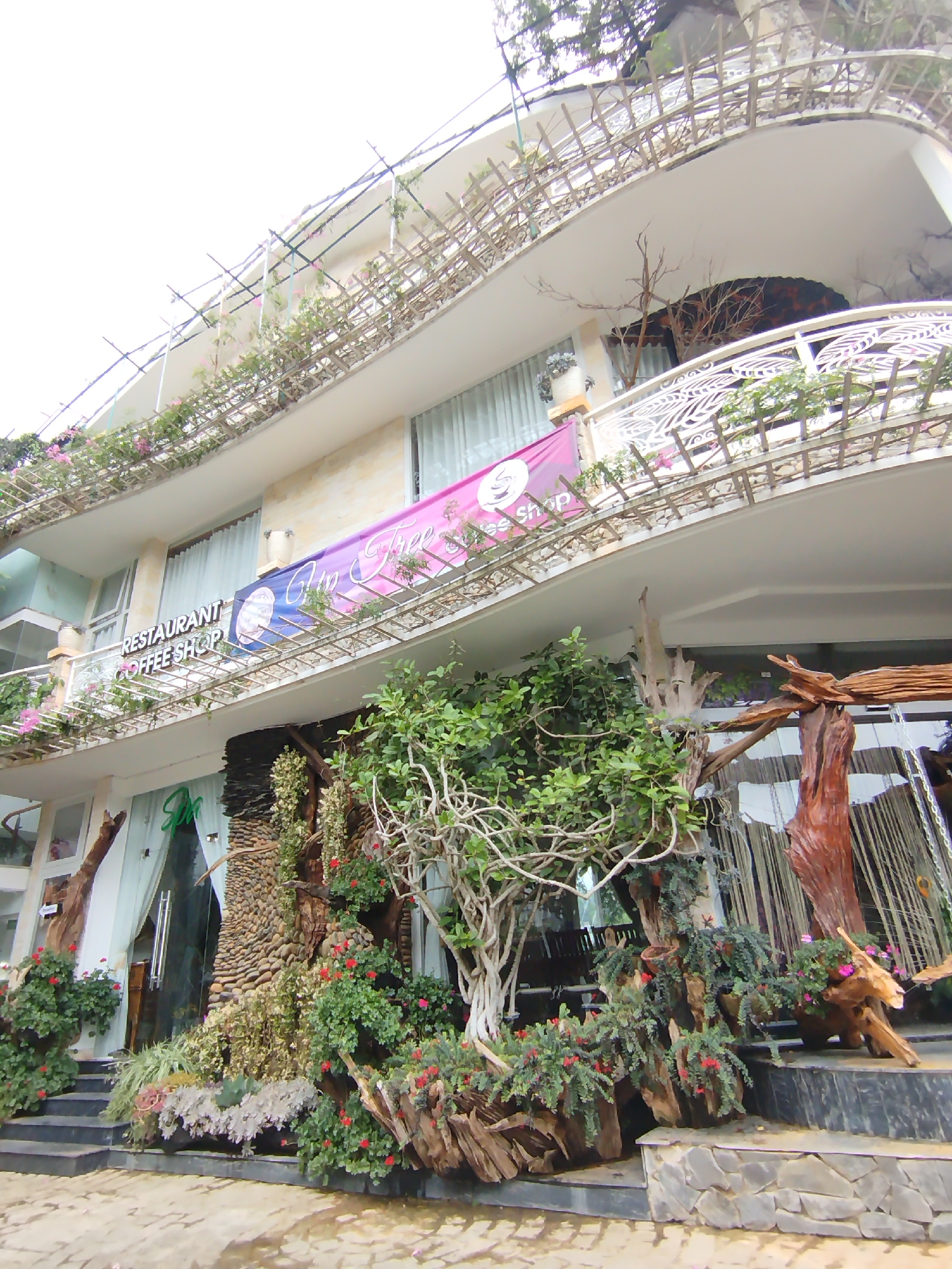The image shows a multi-story building with a facade adorned with plants and decorative elements. The building features curved balconies with ornate railings and bamboo structures, suggesting an outdoor seating or garden area. A banner on one of the balconies reads "RESTAURANT COFFEE SHOP". The ground level is characterized by an entrance with a large glass door, surrounded by lush greenery, stones, and driftwood sculptures. Several stairs lead up to the entrance. The overall impression is of a restaurant or cafe with an emphasis on natural and artistic decor. Based on the architectural style and abundant greenery, this scene is likely in Da Lat, Vietnam, a city known for its French colonial architecture and abundant pine forests and botanical gardens.