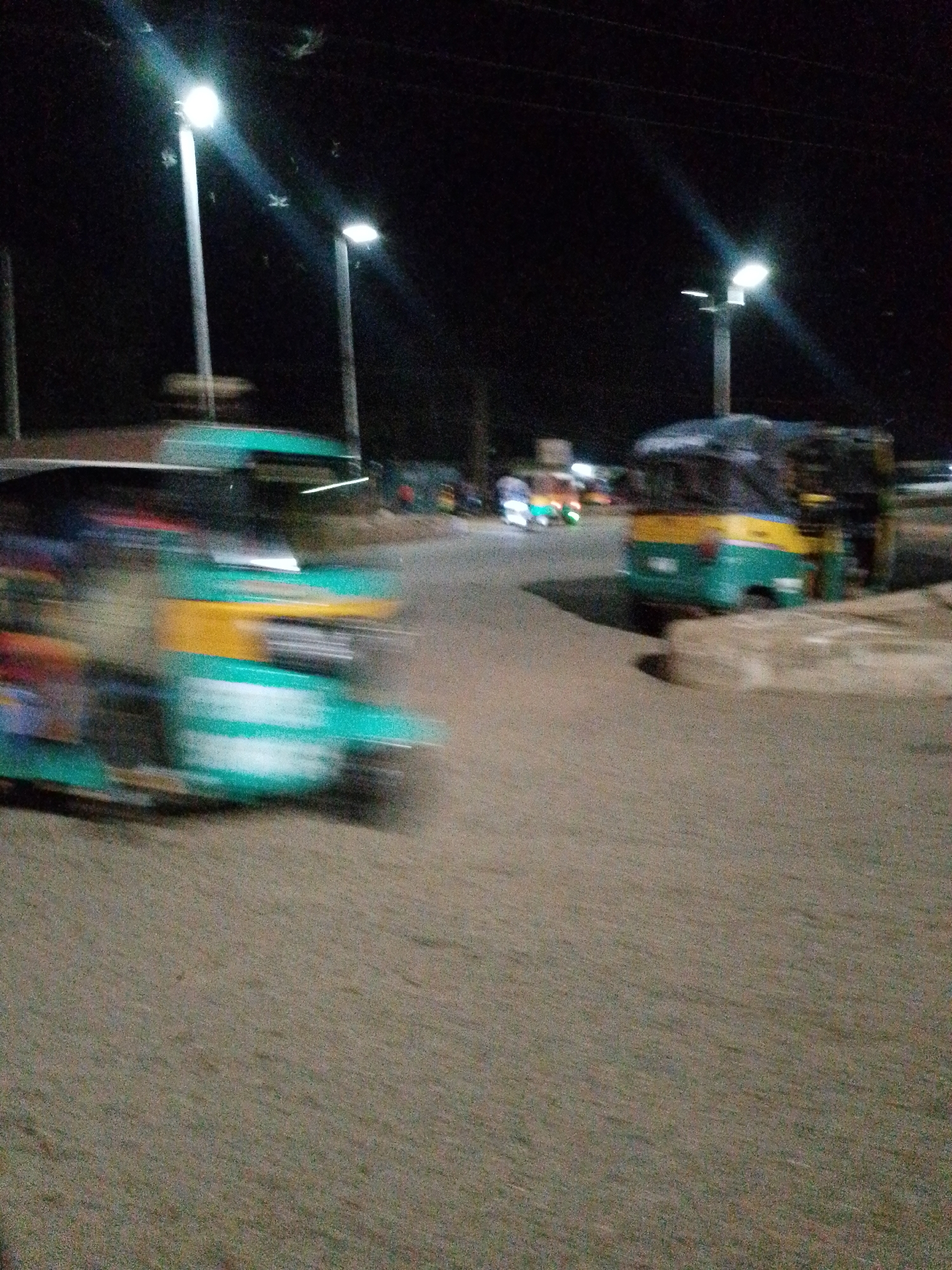 The image depicts a nighttime scene in Bukuru, Nigeria. Streetlights illuminate a road with auto rickshaws (tuk-tuks). One tuk-tuk is in motion, creating a blur, while another is stationary on the right side of the frame. The road surface appears to be paved and is visible in the foreground. The background is dark, with additional tuk-tuks and lights suggesting a populated area.