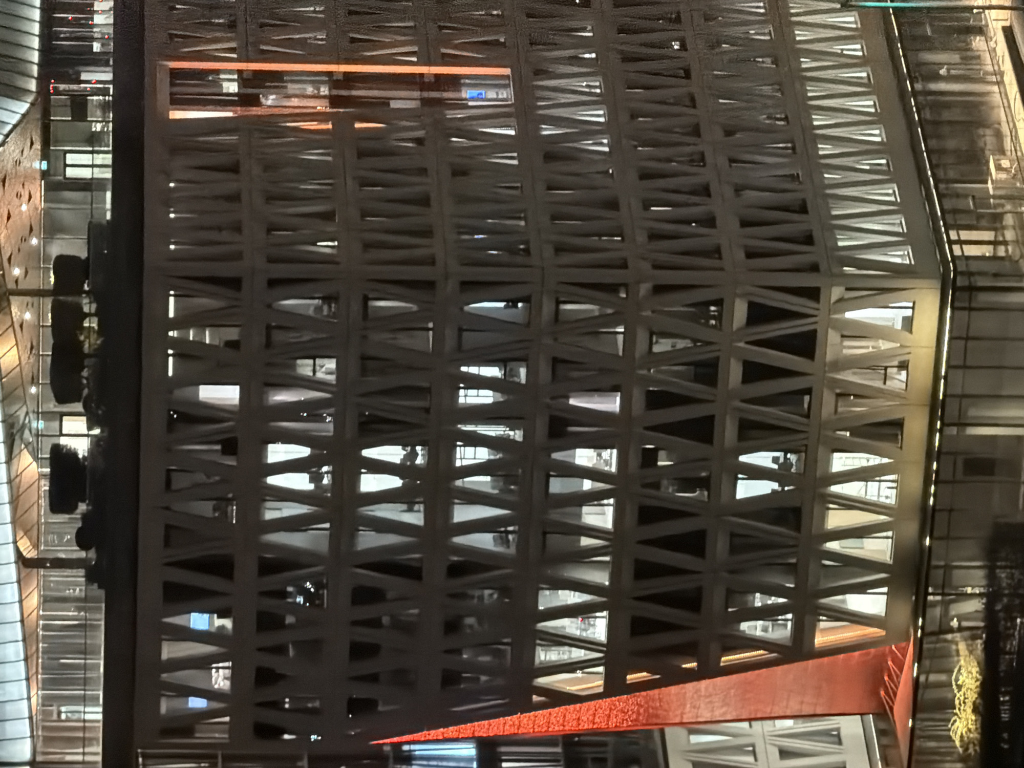The image shows a close-up view of the exterior of a modern building at night, with many windows illuminated. The building has a unique geometric facade with a pattern of intersecting diagonal beams creating triangular openings. A prominent red-colored structure rises along one side of the building, contrasting with the predominantly white and grey tones of the main structure. The lighting from within the building casts a warm glow through the windows, and some areas outside are illuminated by artificial lights, reflecting on a lower level structure. The scene appears to be urban, given the architectural style and lighting.