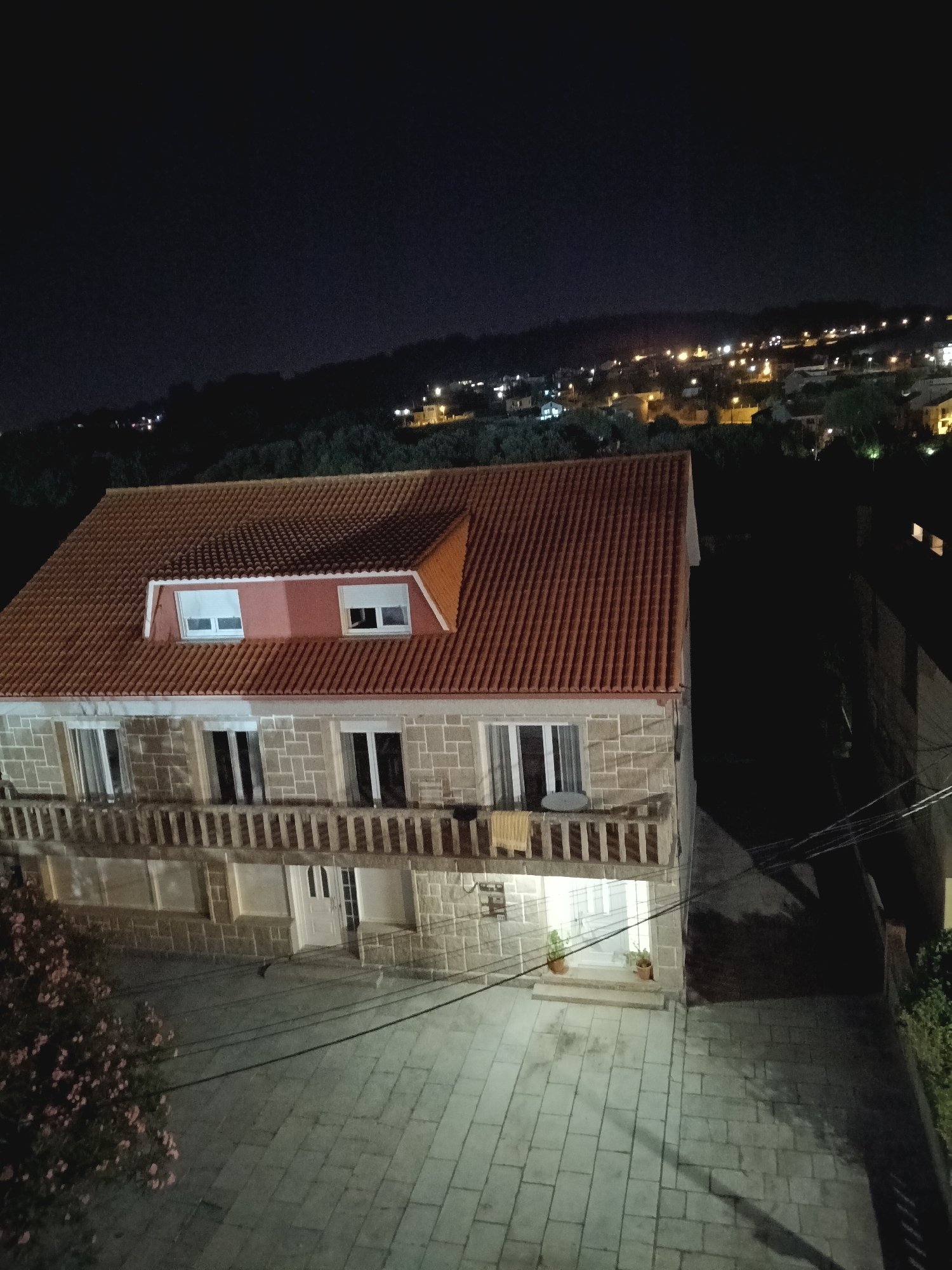 An exterior, high-angle, night shot shows a stone-clad house with a red tiled roof in A Lanzada, Spain. The house has a balcony with a railing on the first floor, with several windows and doors visible on this level and the attic. Two potted plants flank the entrance on the ground floor, which is accessed by a short set of steps. The house is situated in a paved courtyard, with a bush of pink flowers to the left. In the background, a town with illuminated buildings and streetlights is visible on a hillside under a dark sky. Power lines run diagonally across the lower right portion of the image.